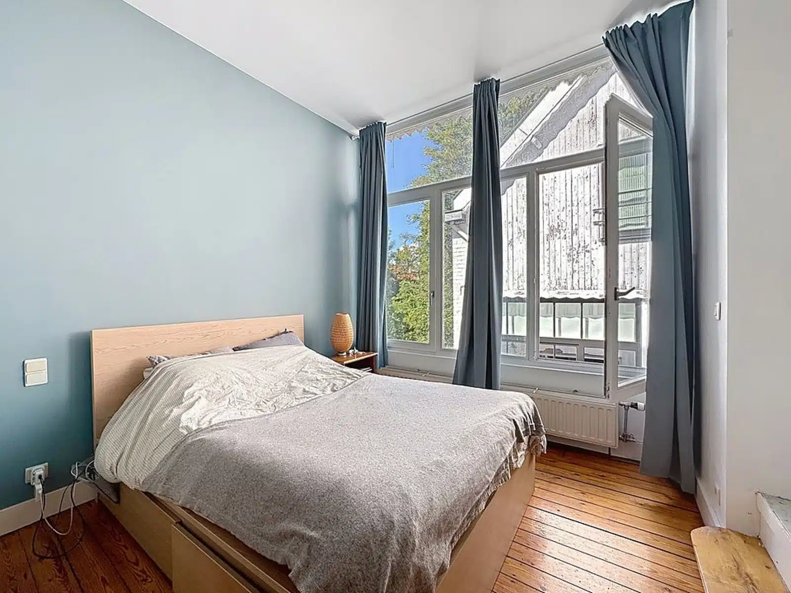 Wijze foto 11 Slaapkamer met blauwe accentmuur, houten bed met grijs beddengoed en een klein nachtkastje met een geweven lamp onder een groot raam met blauwe gordijnen. daglicht stroomt naar binnen vanuit het raam. Radiator zichtbaar onder het raam.