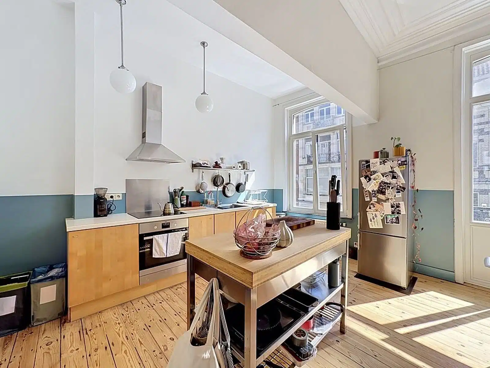 foto 5 Lichte keuken met lichte houten vloeren, een roestvrijstalen eiland en een koelkast bedekt met magneten en foto's achter een grote muur met ramen buiten het gebouw.