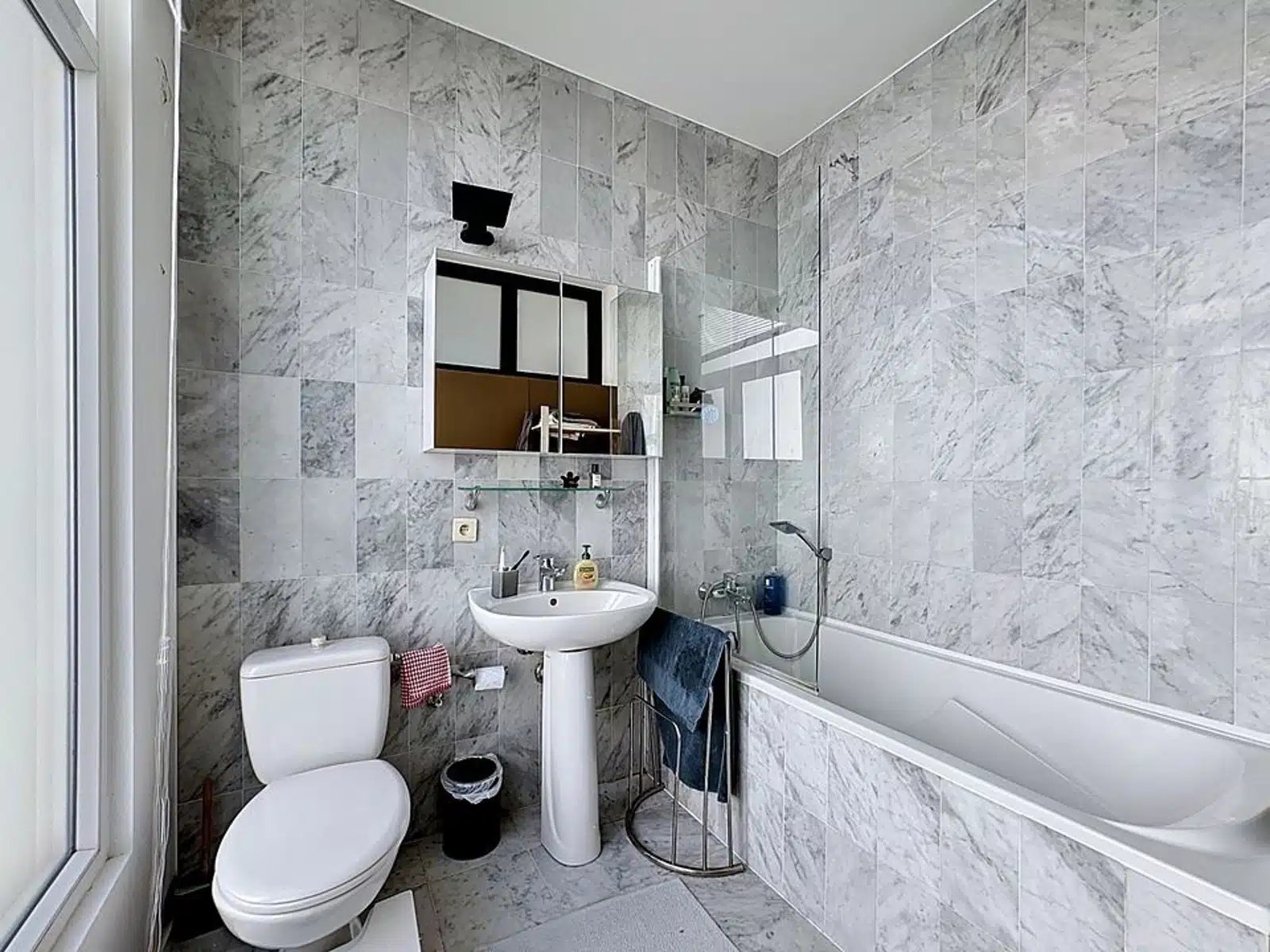 Wijze foto 10 Badkamer met marmeren betegelde muren, wastafel op voet, toilet en een bad/douche combo met glazen paneel; raam aan de linkerkant.