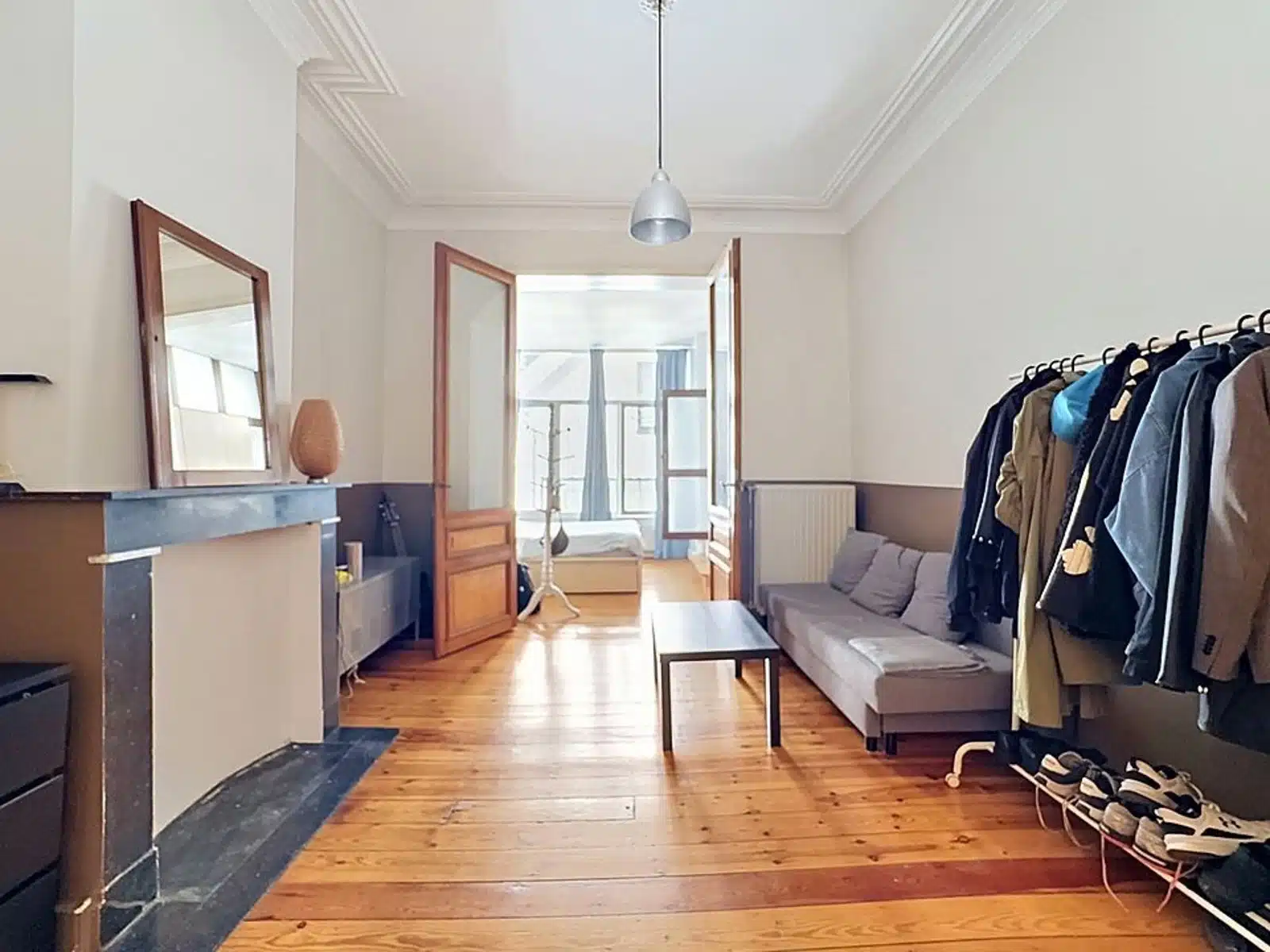 Wijze foto 8 Woonkamer met een kapstok langs de rechtermuur, grijze bank en een salontafel; openslaande deuren leiden naar een lichte aangrenzende kamer met een raam.