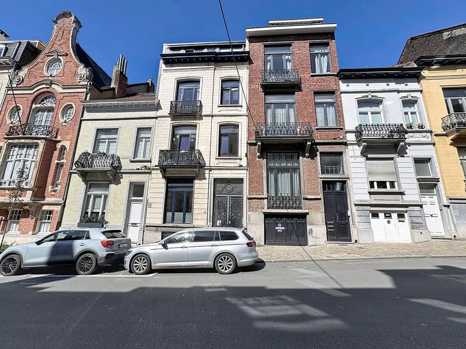 Wijze foto 13 Een rij historische bakstenen en stenen herenhuizen met sierlijke balkons en grote ramen, geparkeerde auto's ervoor in een zonnige straat.