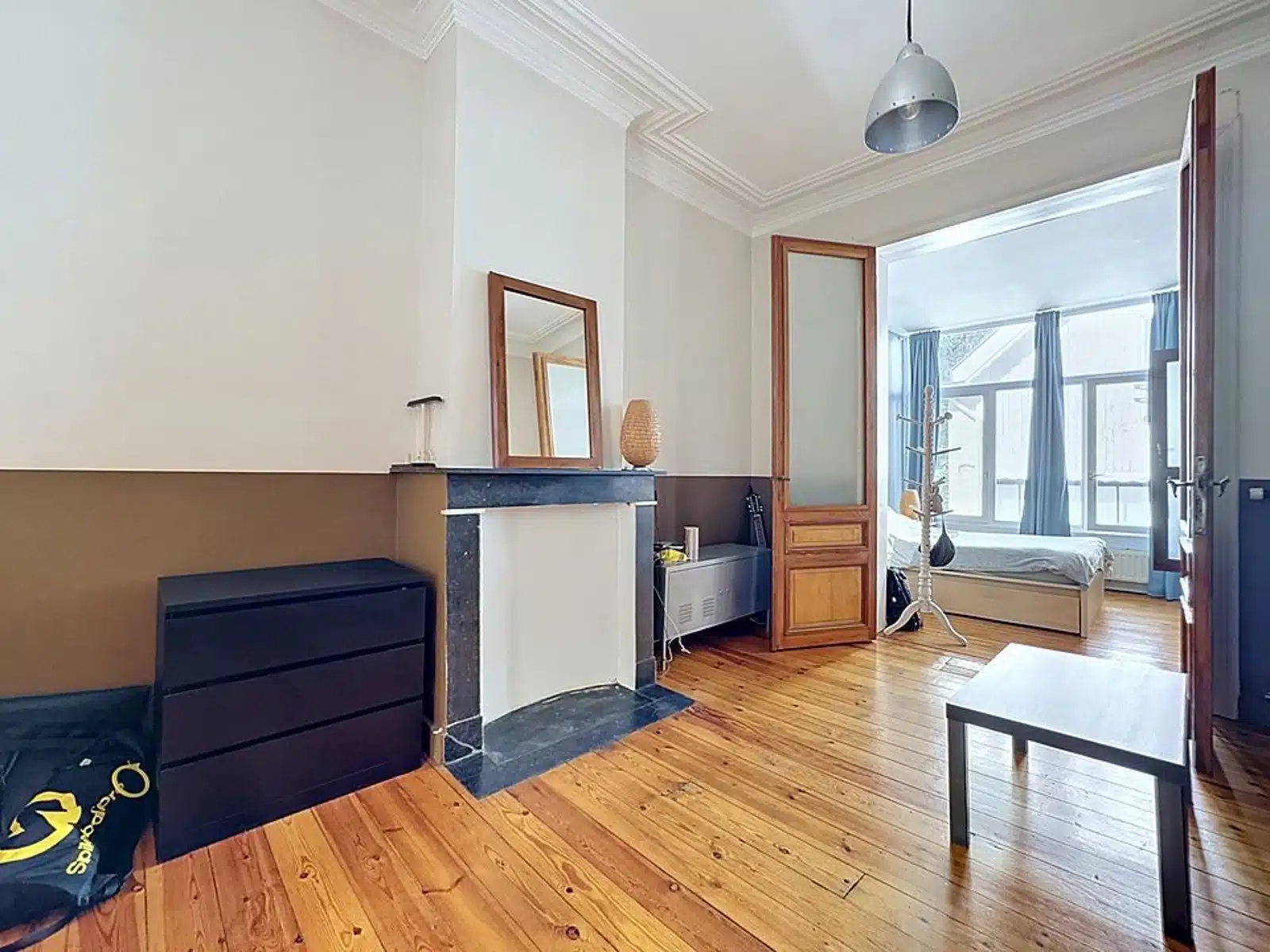 foto 7 Woonkamer met een open haardmantel en spiegel, open deur die leidt naar een lichte slaapkamer met een bed en grote raamstijlen met blauwe gordijnen zichtbaar aan het uiteinde.