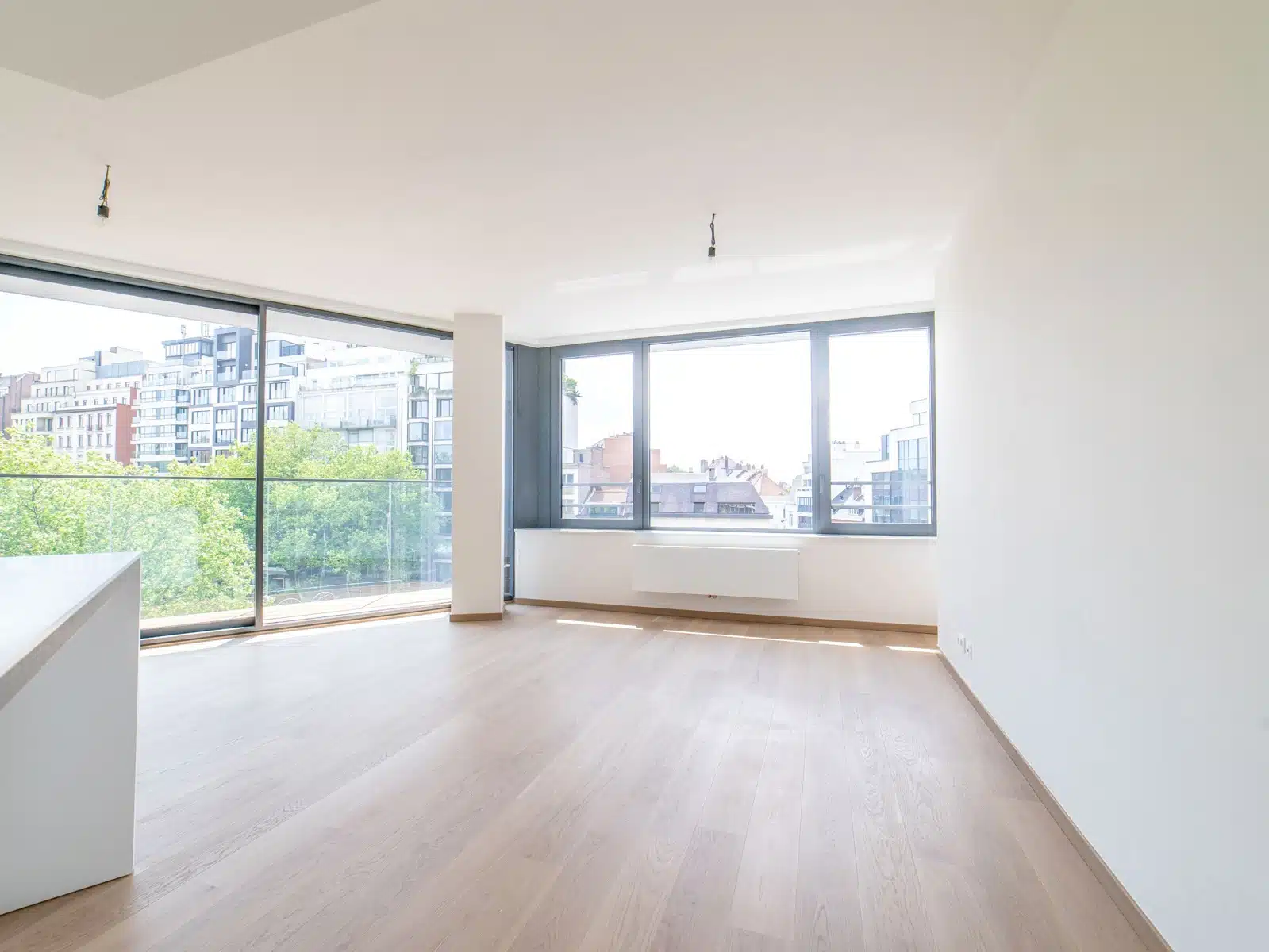 whise photo 6 Bright empty living room with large floor-to-ceiling windows overlooking a cityscape and trees, light wood flooring.