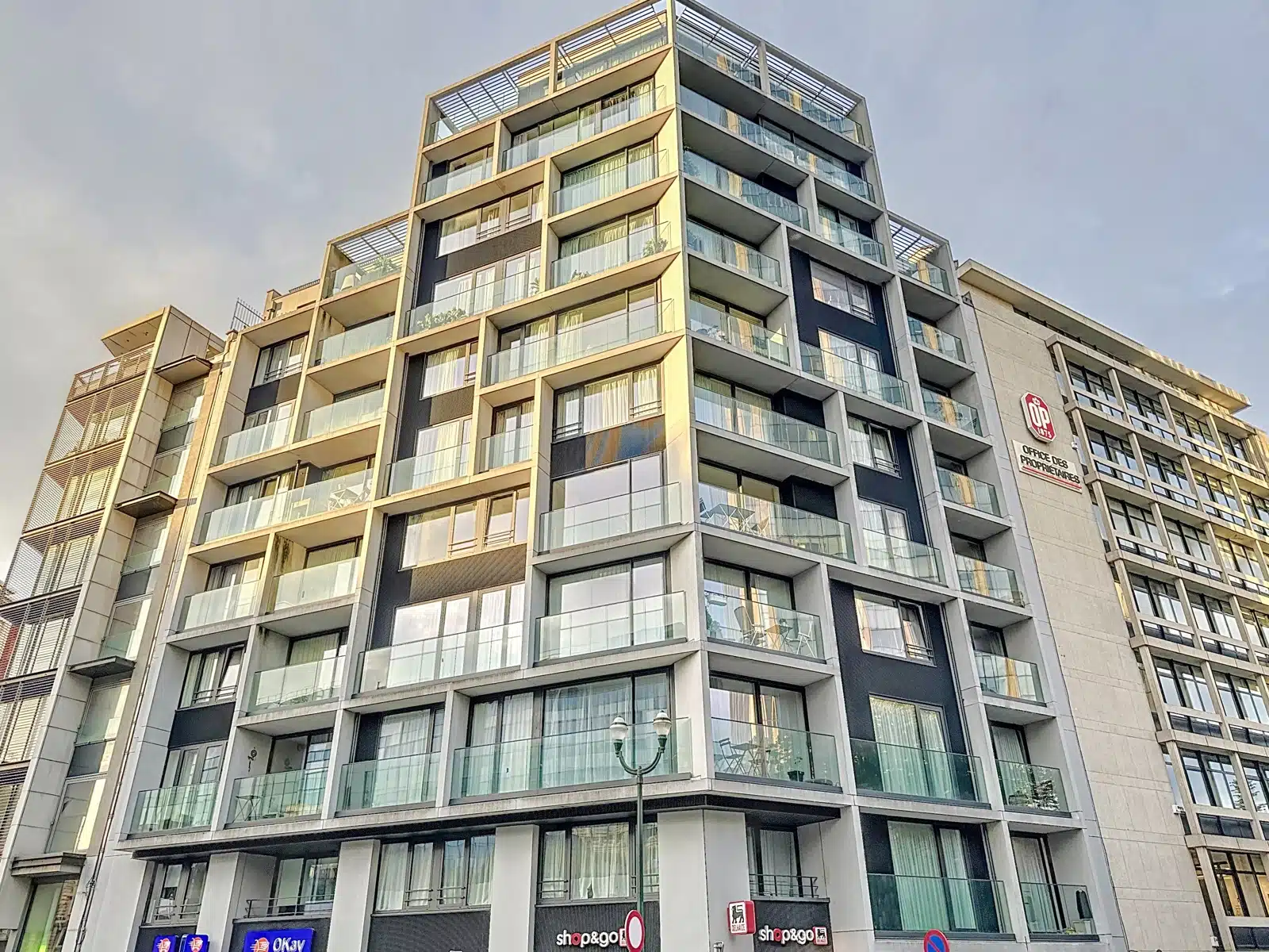 Façade Moderne Avec Balcons Vitrés – You Real Estate Agency Immeuble moderne avec appartements, balcons vitrés et commerces en rez-de-chaussée.