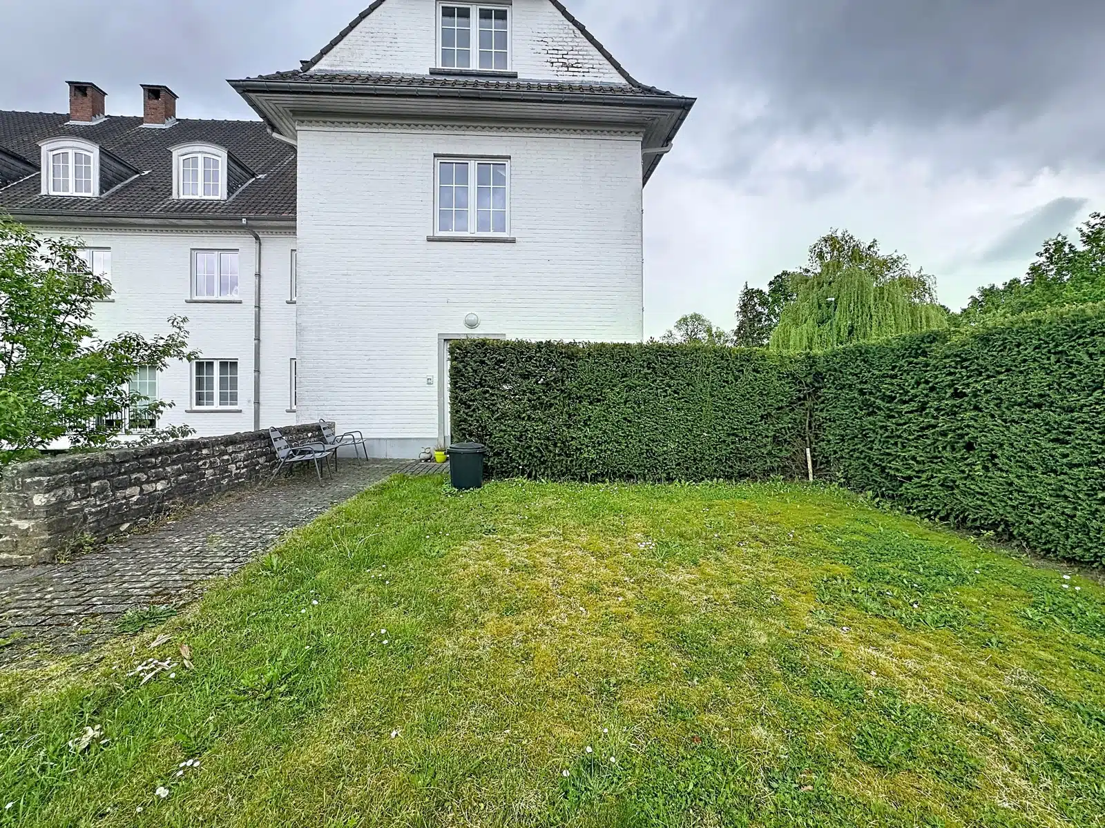 whise photo 9 White brick building with a pitched roof, small paved courtyard, and metal benches along a low stone wall; a tall green hedge encloses the yard under a cloudy sky.