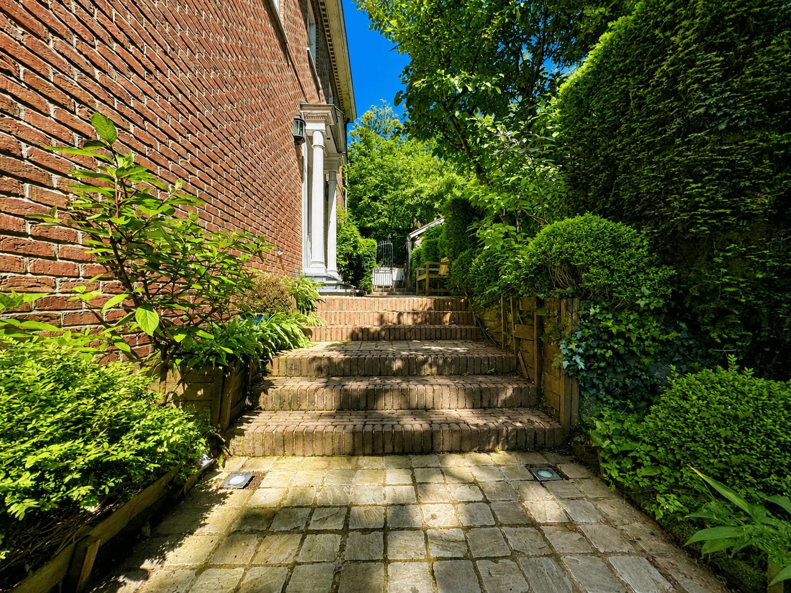 Entrée Latérale avec Jardin Arboré – You Real Estate Agency Terrasse pavée et entrée latérale d’une maison en briques avec jardin arboré.