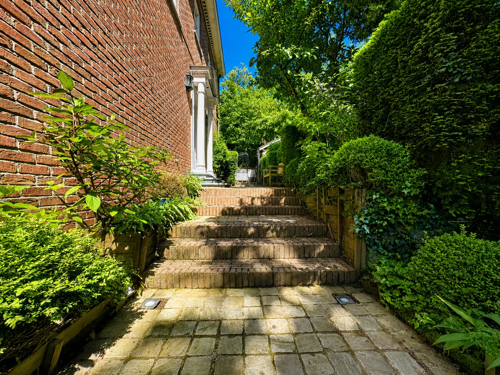 Entrée Latérale avec Jardin Arboré – You Real Estate Agency Terrasse pavée et entrée latérale d’une maison en briques avec jardin arboré.