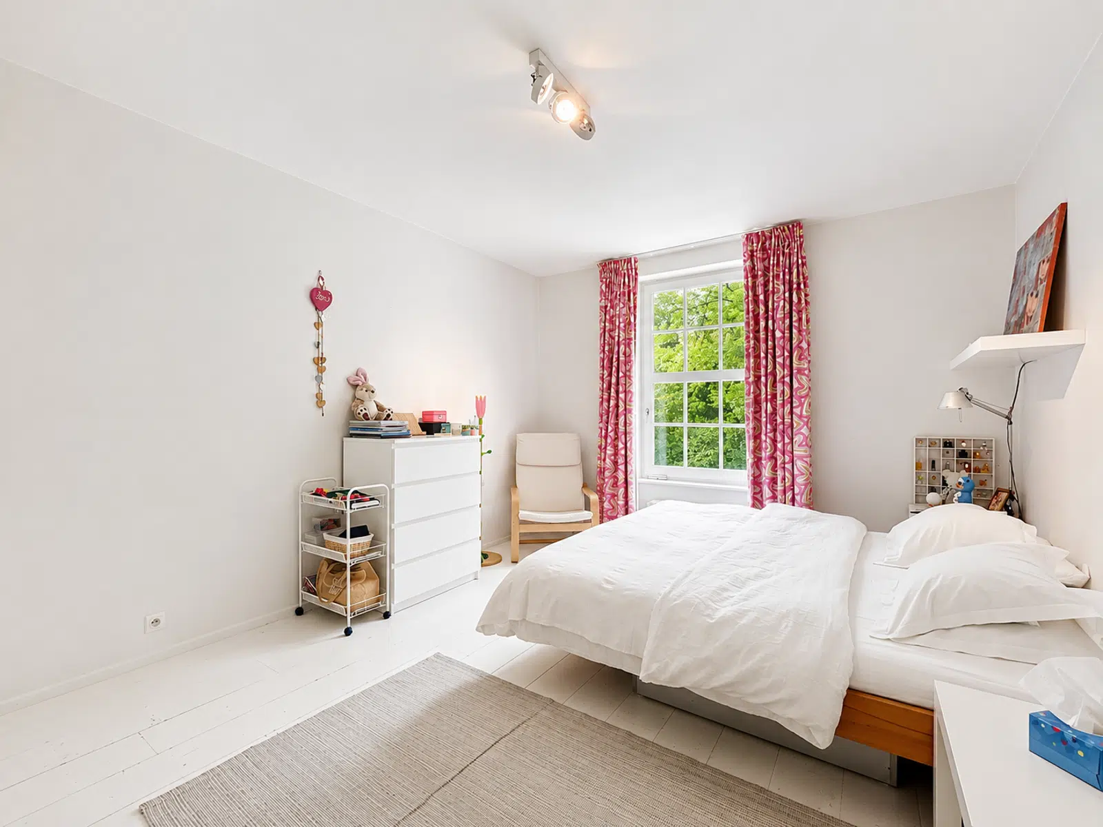 Chambre Lumineuse avec Vue Verdoyante – You Real Estate Agency Chambre lumineuse d’appartement avec lit blanc, fenêtre sur verdure et rideaux roses.