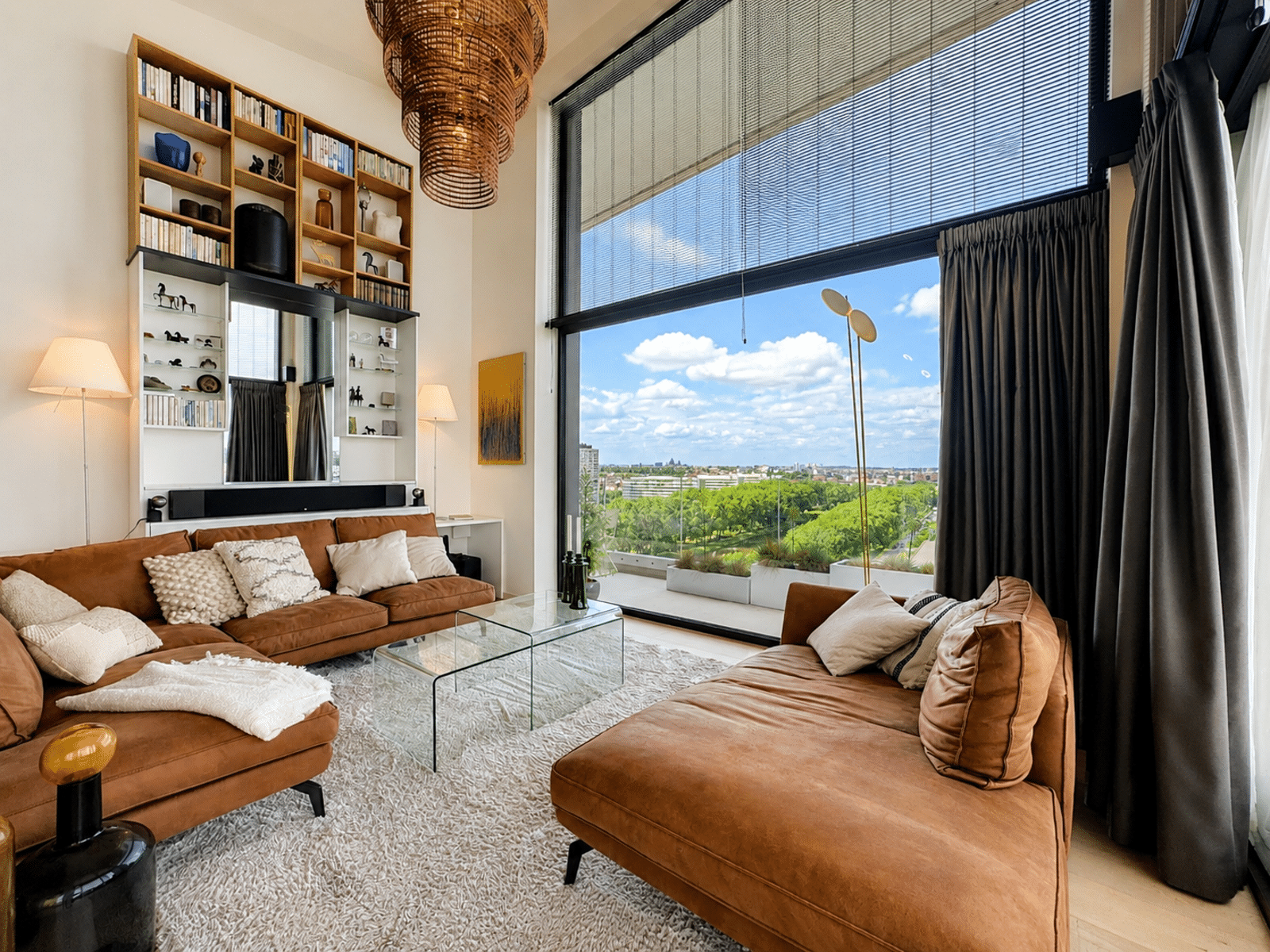 whise photo 6 Living room with a brown suede sectional, glass coffee table, and a tall bookcase along the wall, large window showing a cityscape outside.