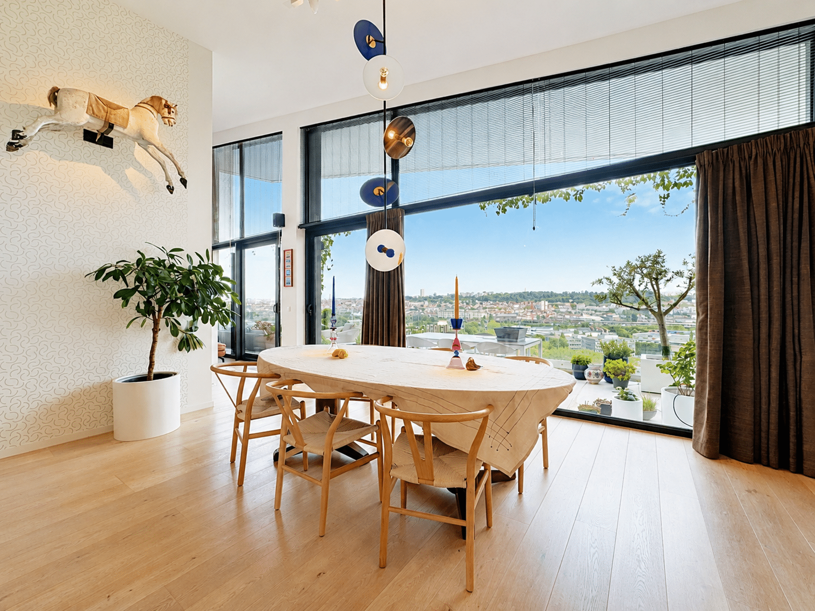 whise photo 7 Bright dining room with oval wooden table, four chairs, large windows showing a city view, a potted plant, and a mounted horse sculpture on patterned wallpaper wall.