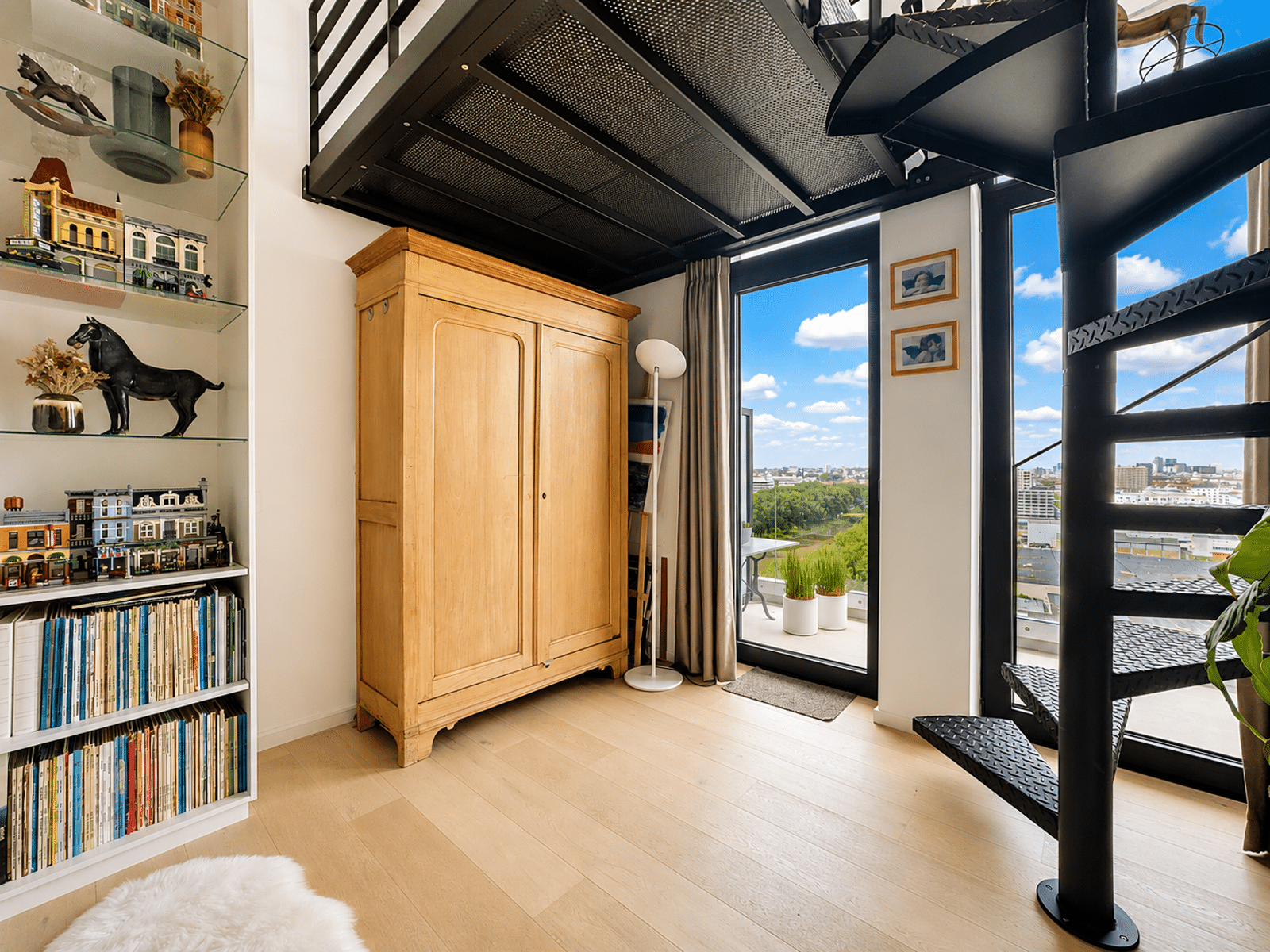 Salon Loft Lumineux avec Balcon – You Real Estate Agency Salon d’appartement loft avec mezzanine, escalier métallique, balcon et vue urbaine.