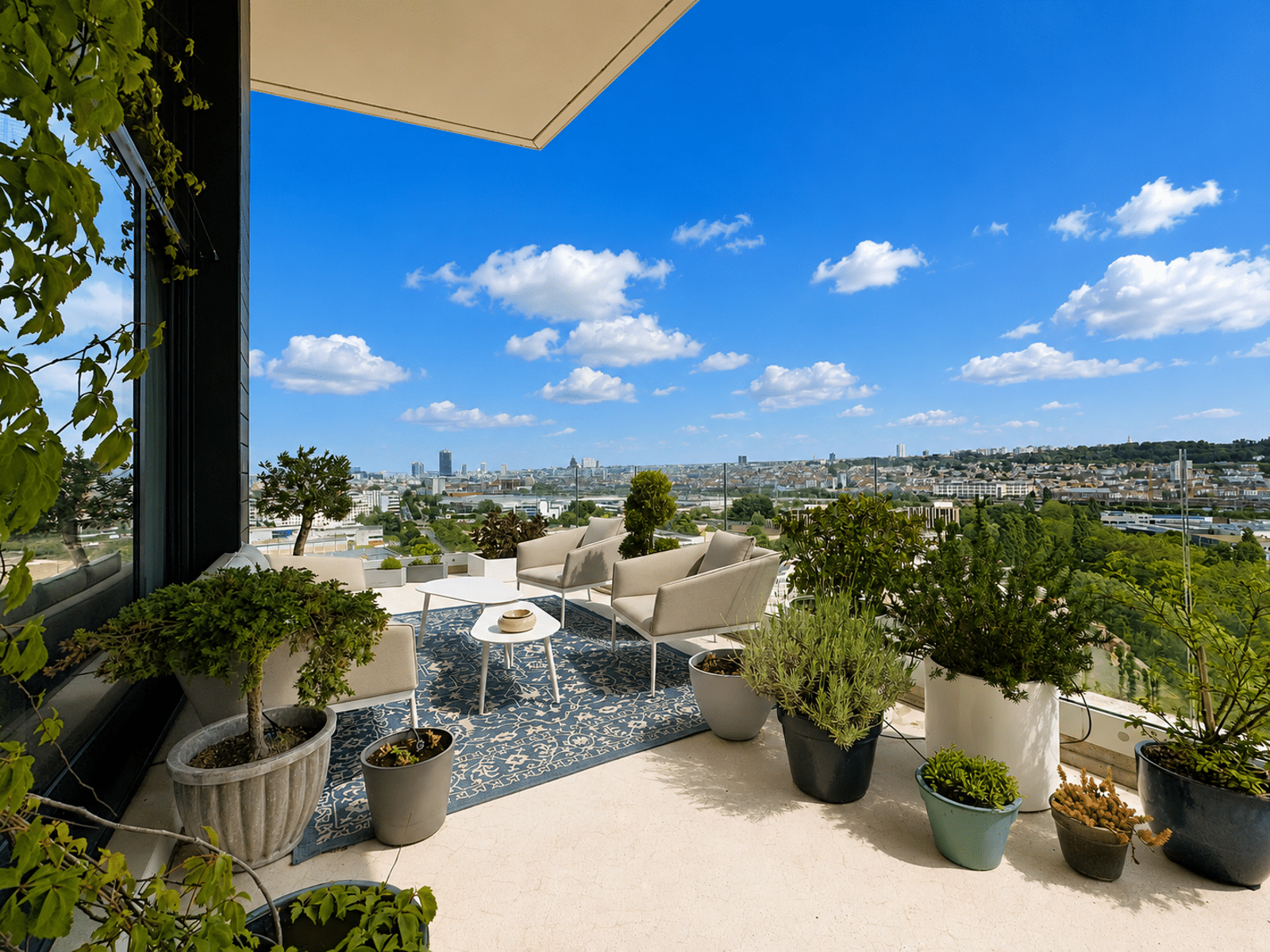 Terrasse avec Vue sur la Ville – You Real Estate Agency Terrasse d’appartement moderne avec salon extérieur, plantes et vue panoramique sur la ville.