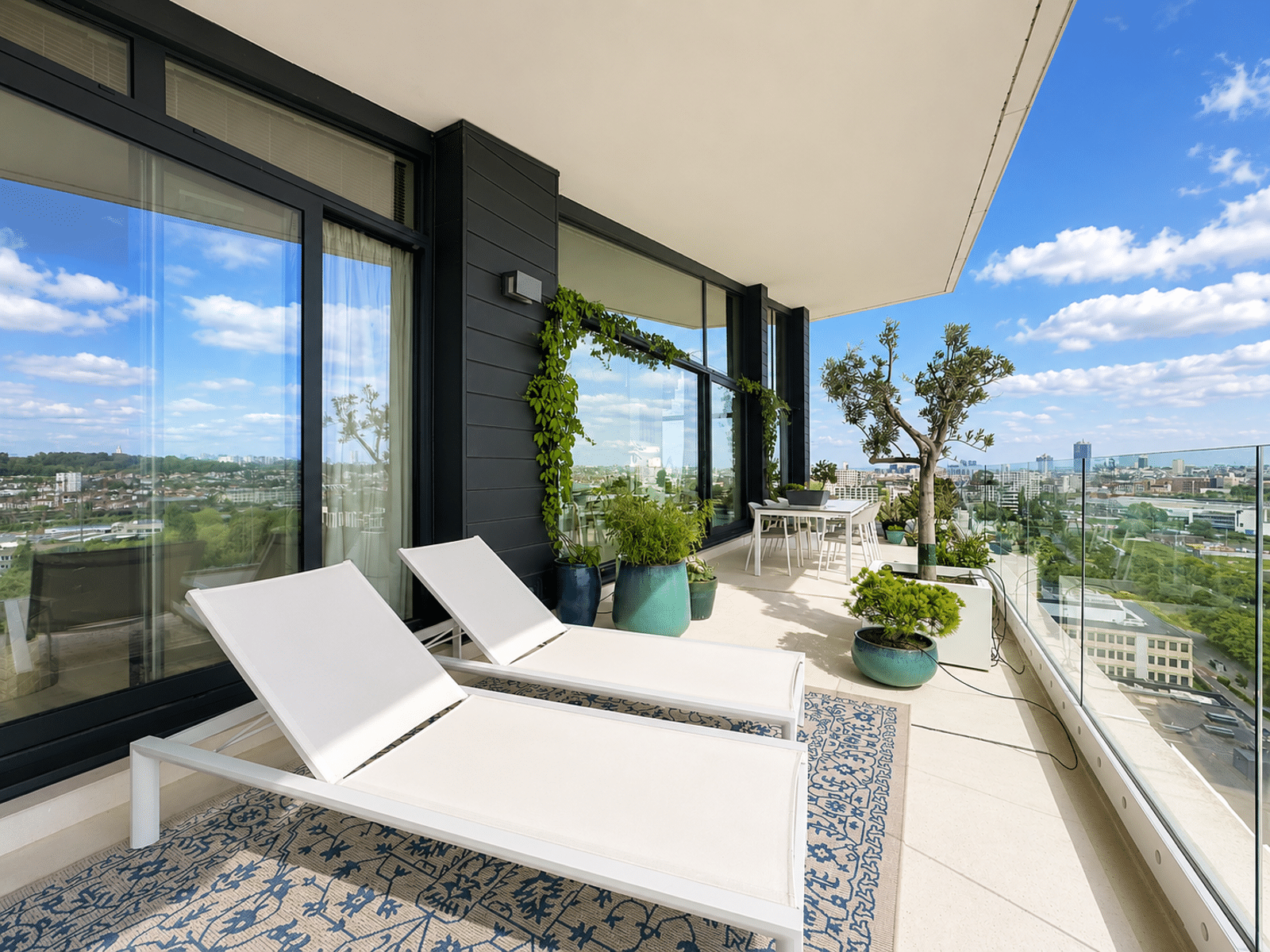 whise photo 3 Modern balcony with glass railing, white lounge chairs, and potted plants overlooking a city skyline on a sunny day.