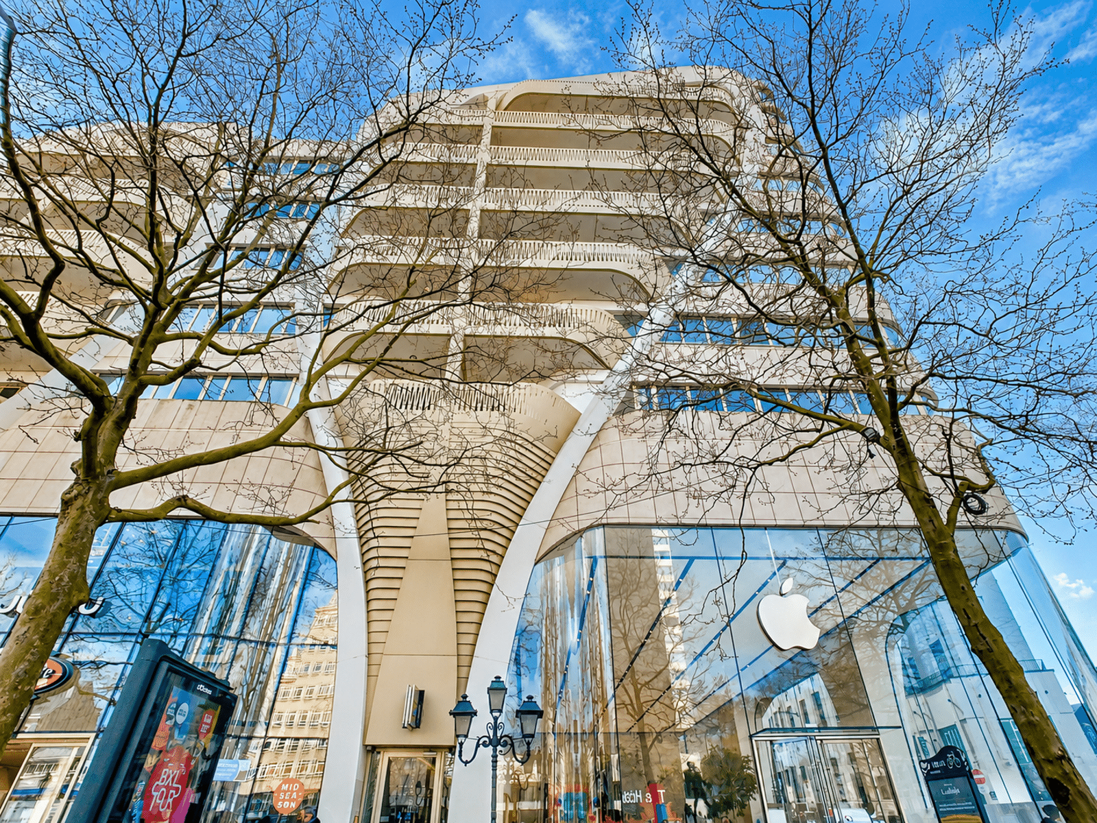 whise photo 11 Modern multi-story building with curved balconies and a glass storefront, framed by leafless trees against a blue sky (Apple store visible).