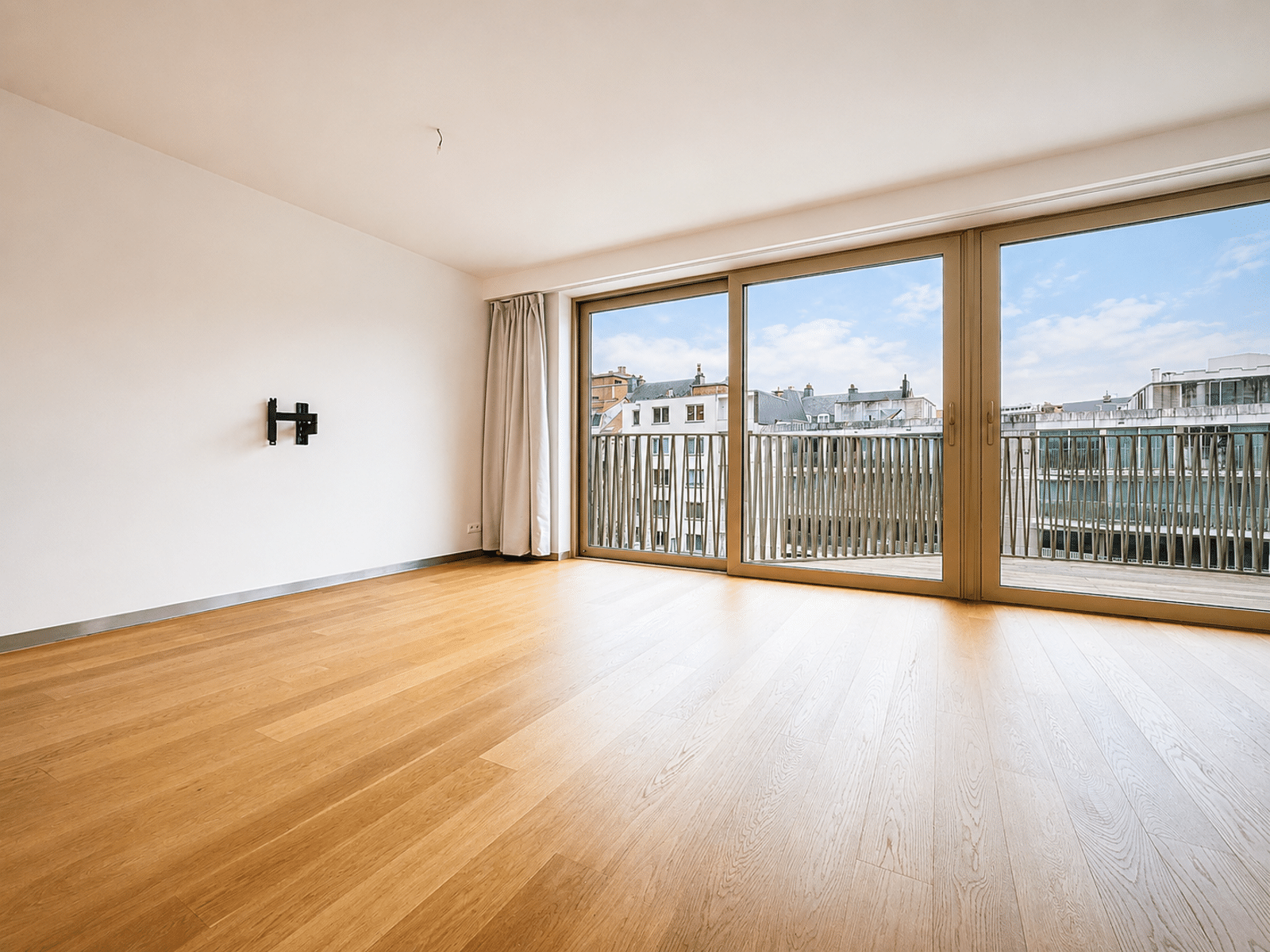 Salon Lumineux avec Balcon – You Real Estate Agency Bright, empty living room with a hardwood floor and large glass doors opening to a balcony overlooking city rooftops.