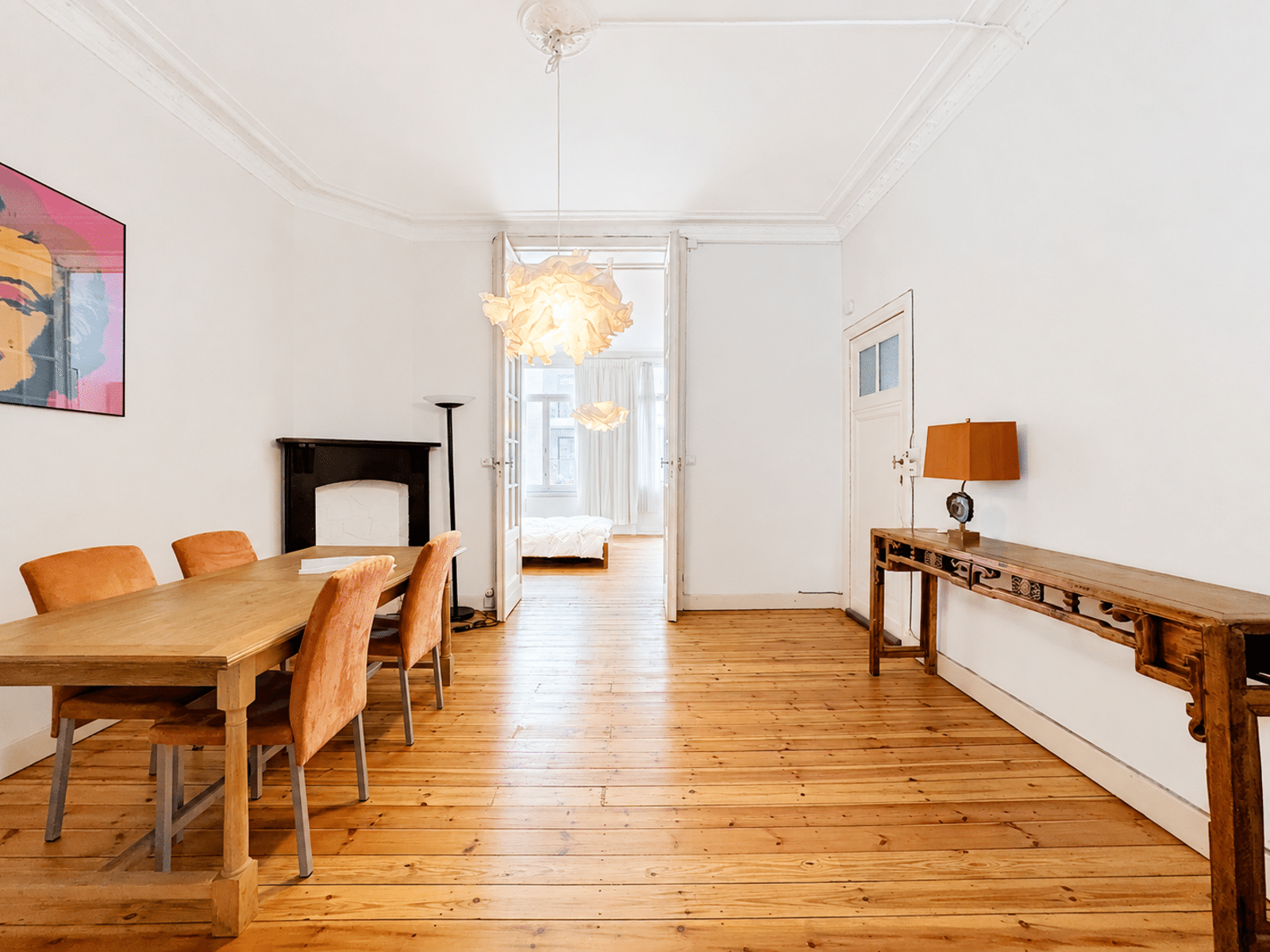 foto 3 Eetkamer met een houten tafel en zes oranje stoelen, een zwarte open haard en een bewerkte houten consoletafel met een oranje lampenkap; dubbele deuren openen naar een lichte slaapkamer.