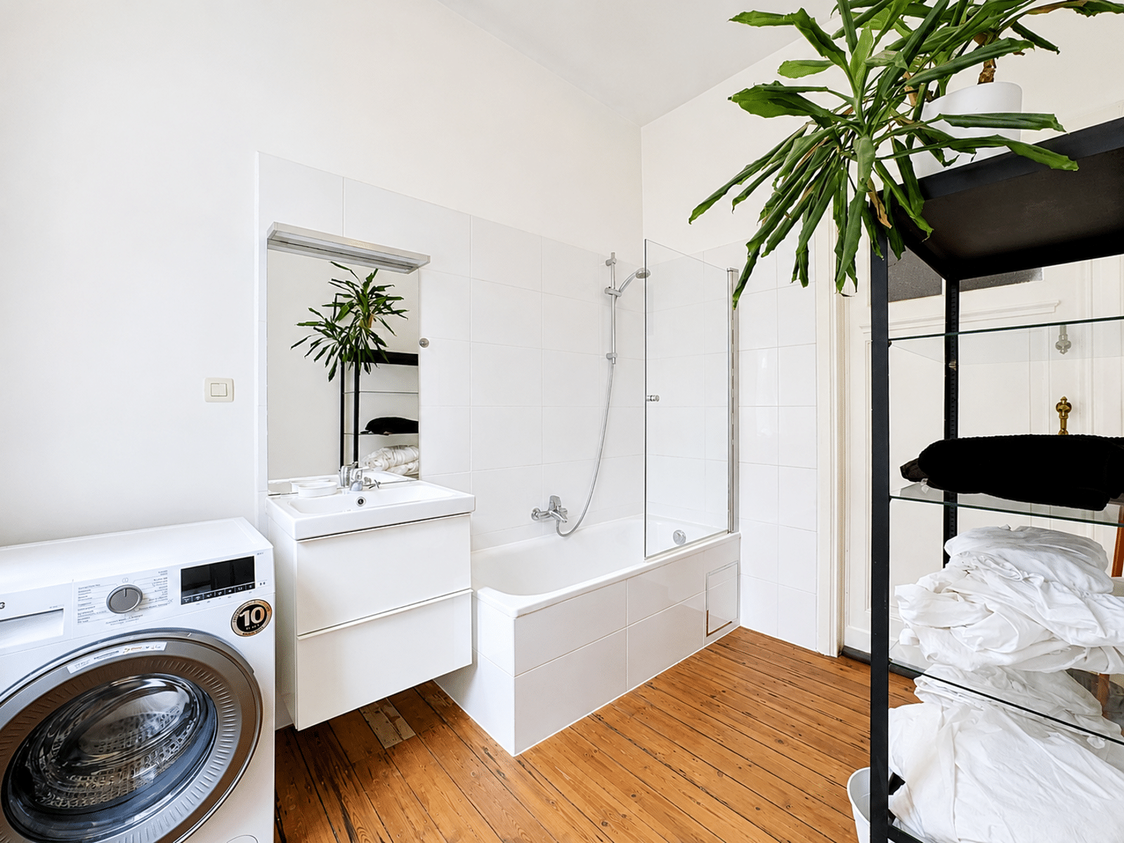 Wijze foto 8 Lichte badkamer met een wasmachine, wit badmeubel en een bad met glazen scherm; in de hoek staat een grote groene plant.