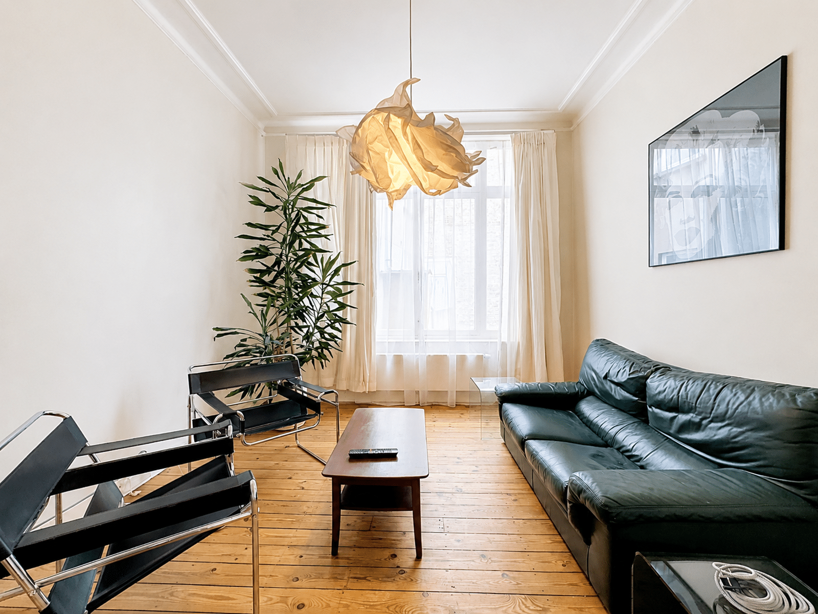 foto 6 Lichte minimalistische woonkamer met een groene leren bank, houten salontafel en een sculpturale hanglamp. Een hoge plant staat in de hoek bij een groot raam met transparante gordijnen.