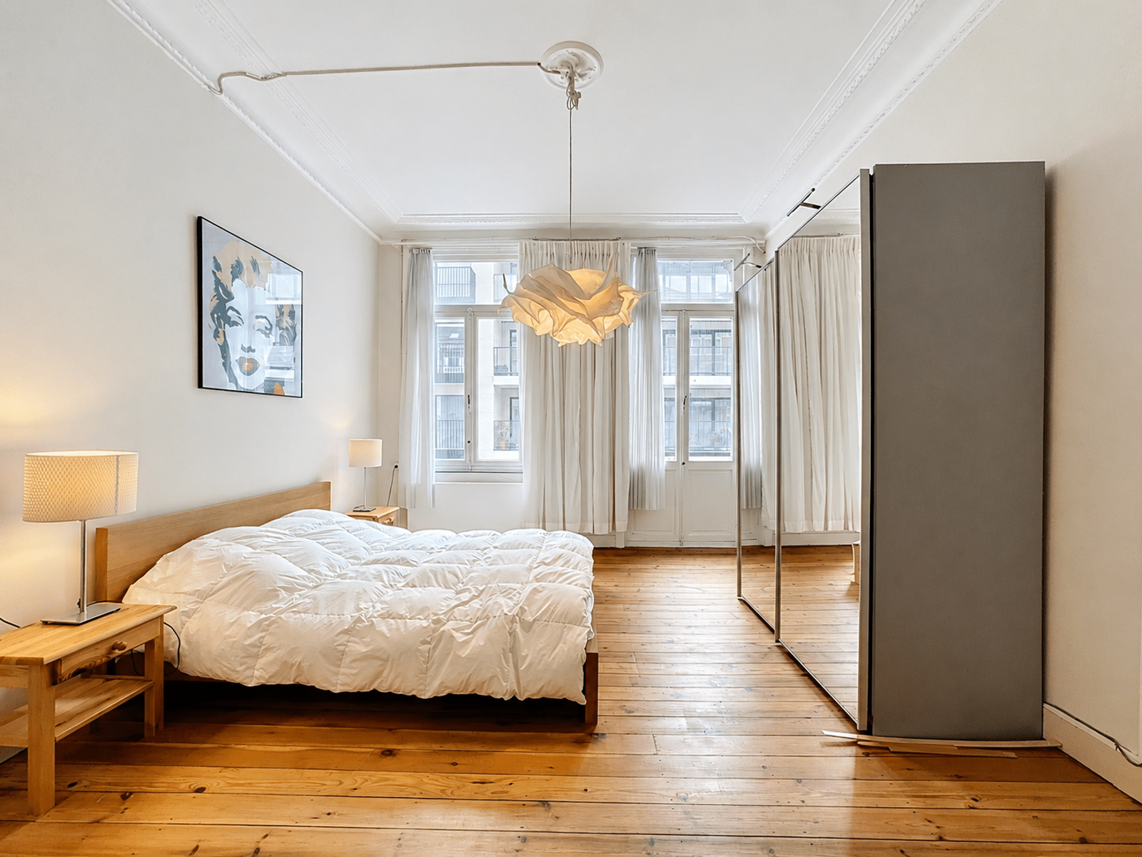 foto 7 Lichte moderne slaapkamer met wit beddengoed, houten vloeren, een ingelijste kunstprint, twee bedlampjes en een grote kledingkast met spiegeldeuren.