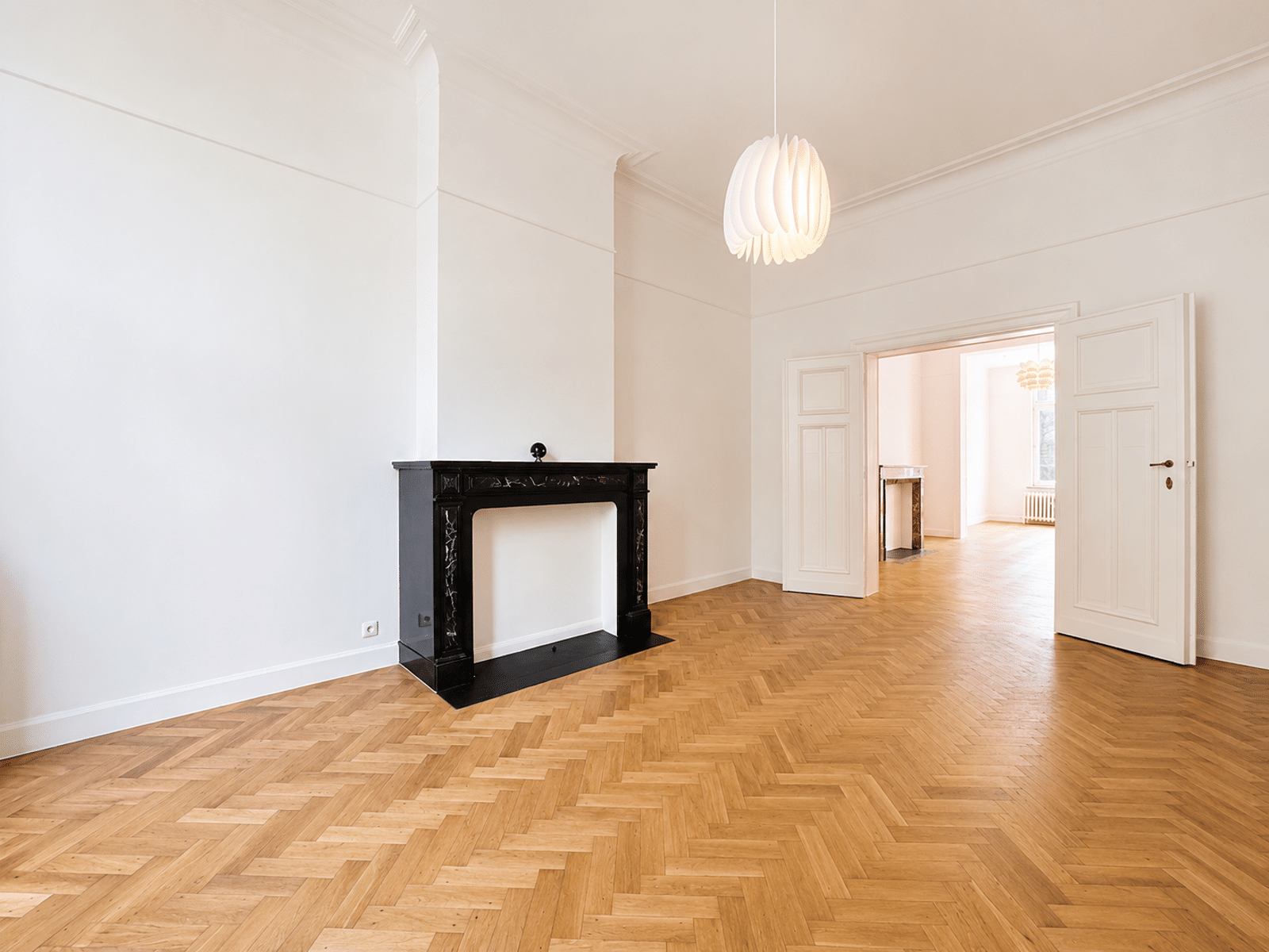 Bright living room with fireplace - You Real Estate Agency Salon d’appartement lumineux avec parquet en point de Hongrie, cheminée noire et double porte.