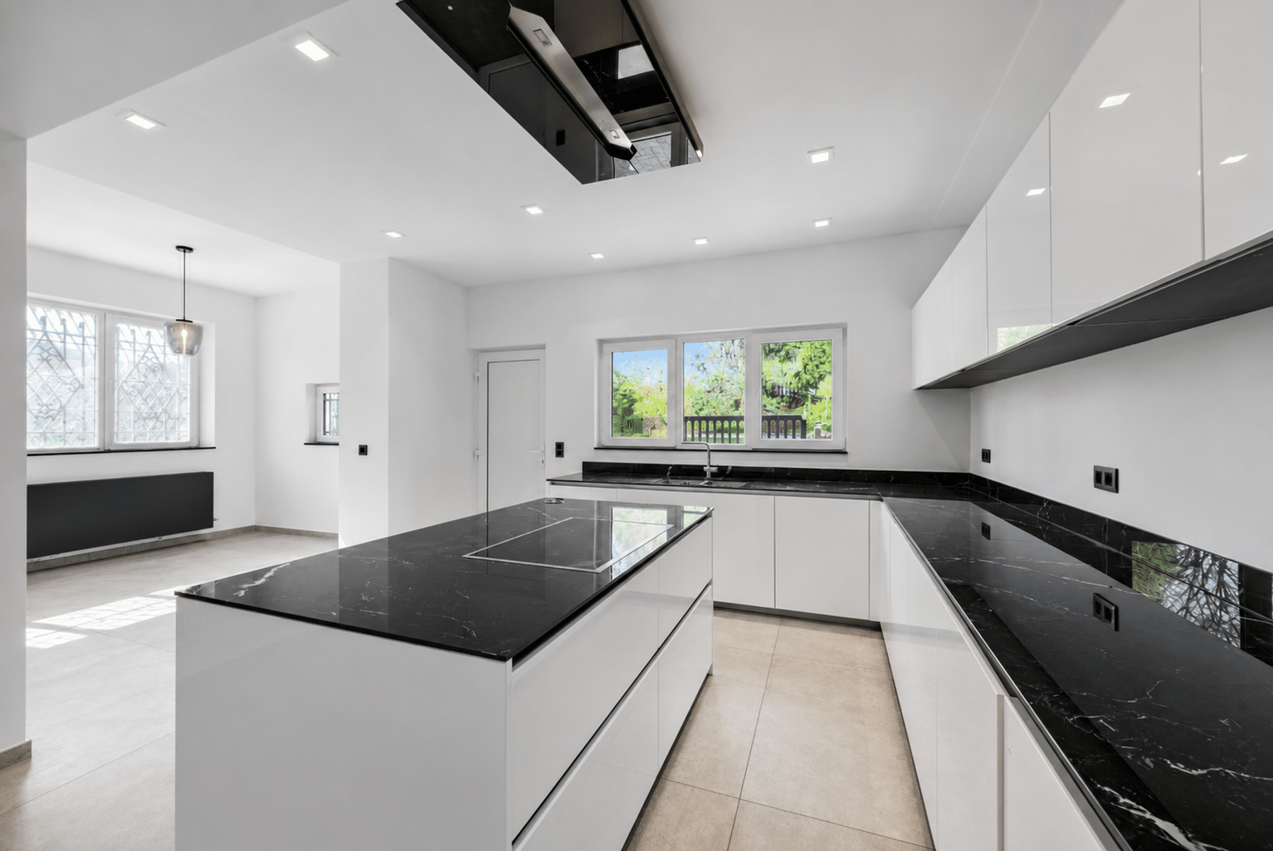 Wijze foto 8 Moderne witte keuken met zwart marmeren aanrechtblad en een groot eiland, verlicht door plafondverlichting en met uitzicht op een tuin via grote ramen achter het aanrecht.