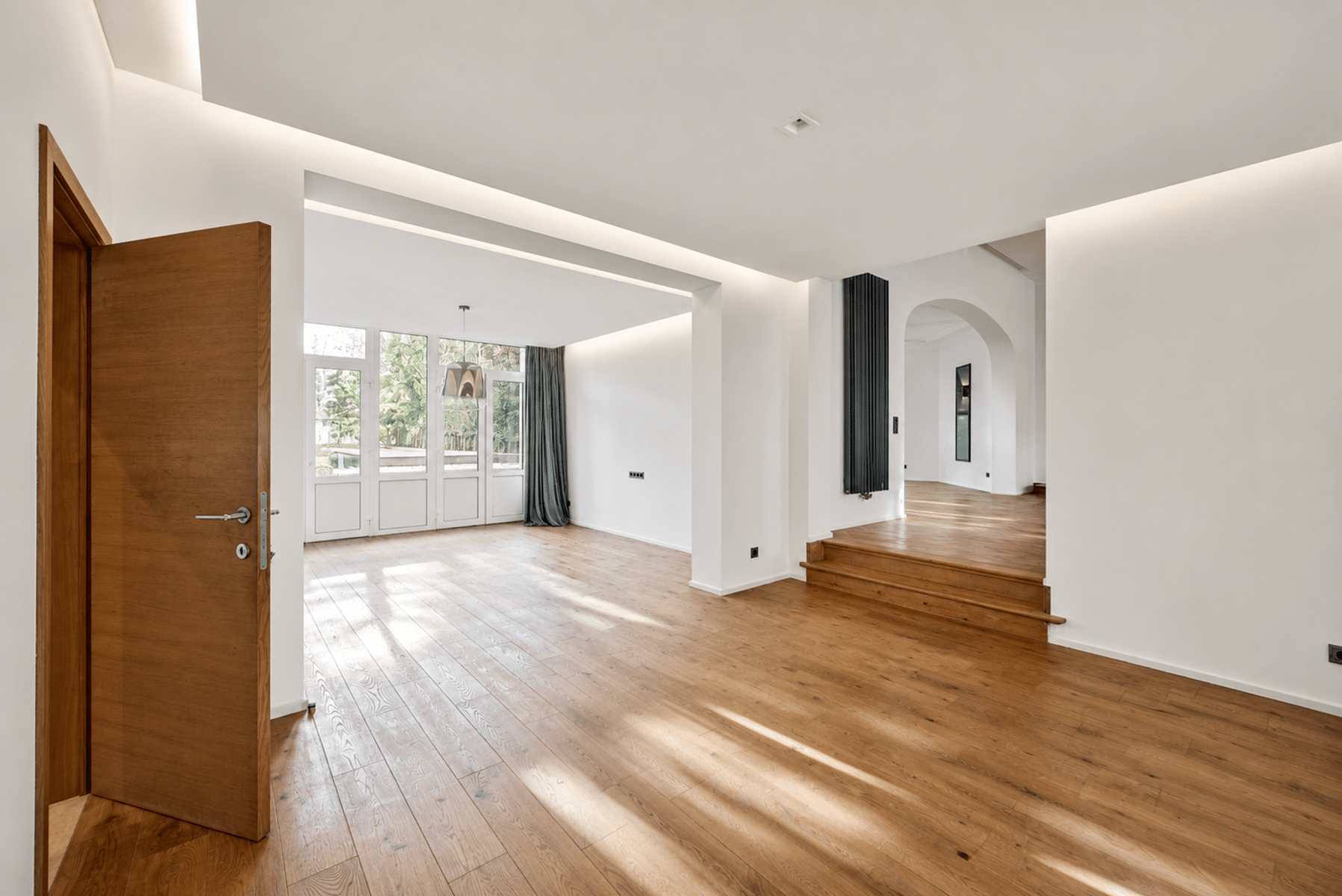 Wijze foto 9 Open, zonovergoten woonkamer met warme houten vloeren, witte muren en een grote glazen wand die daglicht binnenlaat; links staat een houten deur op een kier.
