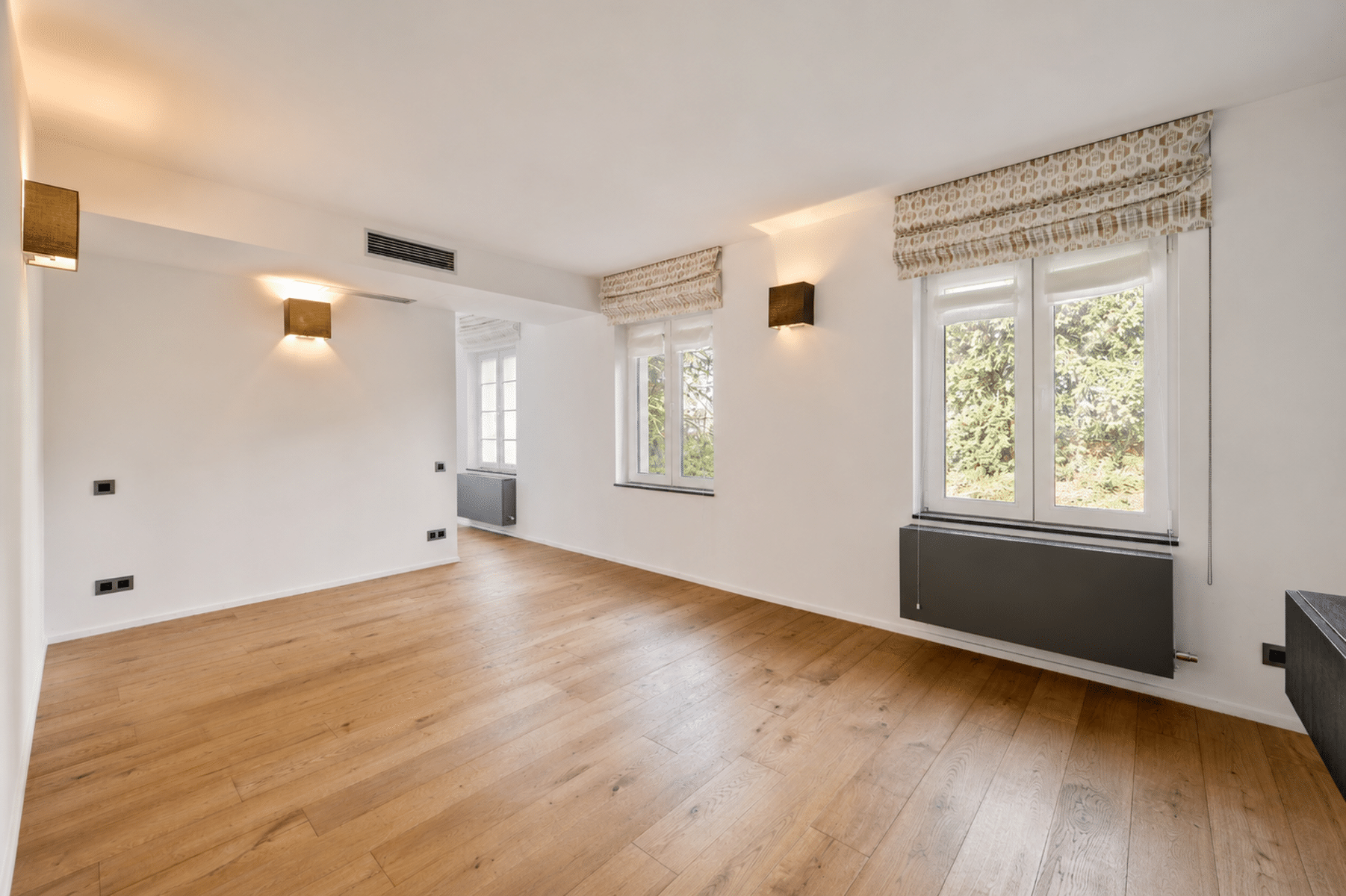 Lichte woonkamer met parketvloer - You Real Estate Agency Lichte, lege woonkamer met parketvloer, grote ramen en witte muren.