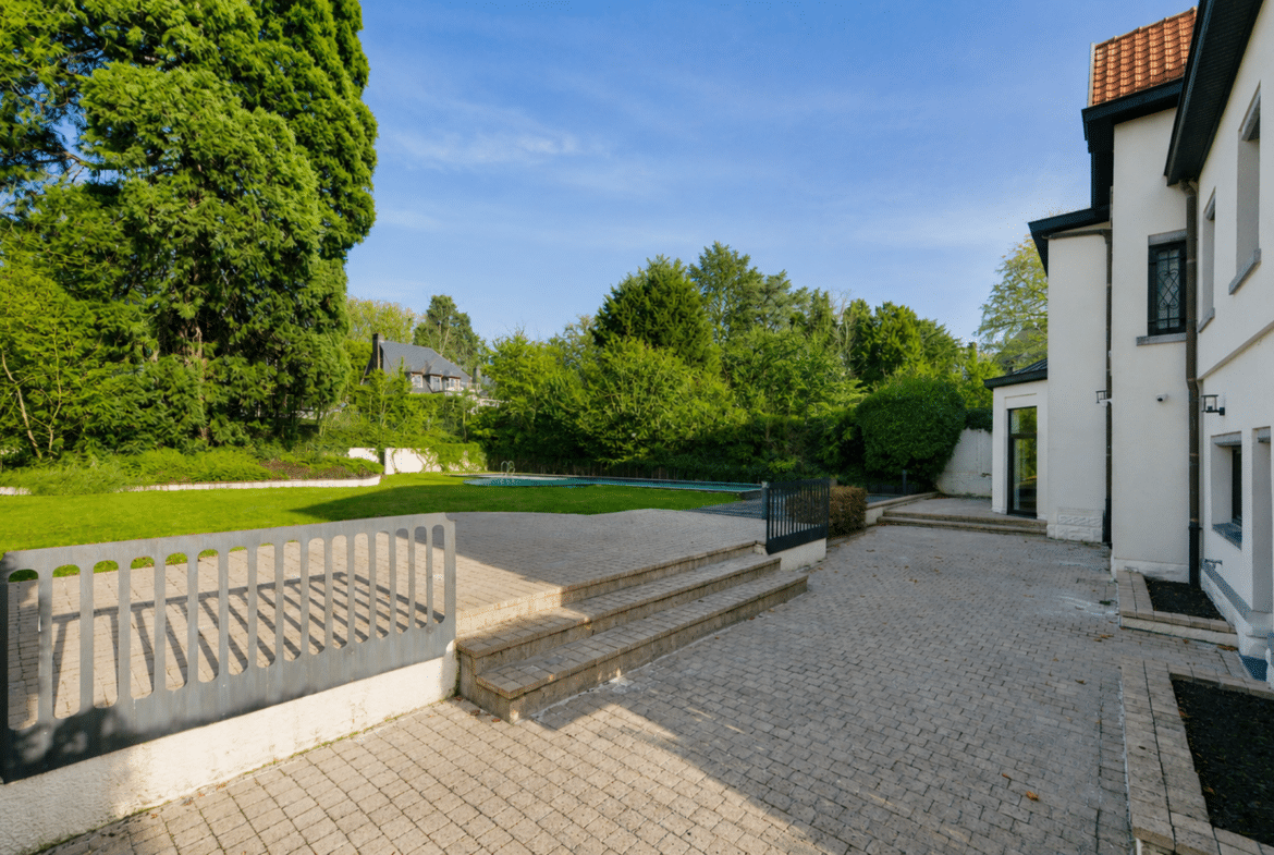 Maison blanche avec grande terrasse pavée, piscine et jardin arboré.