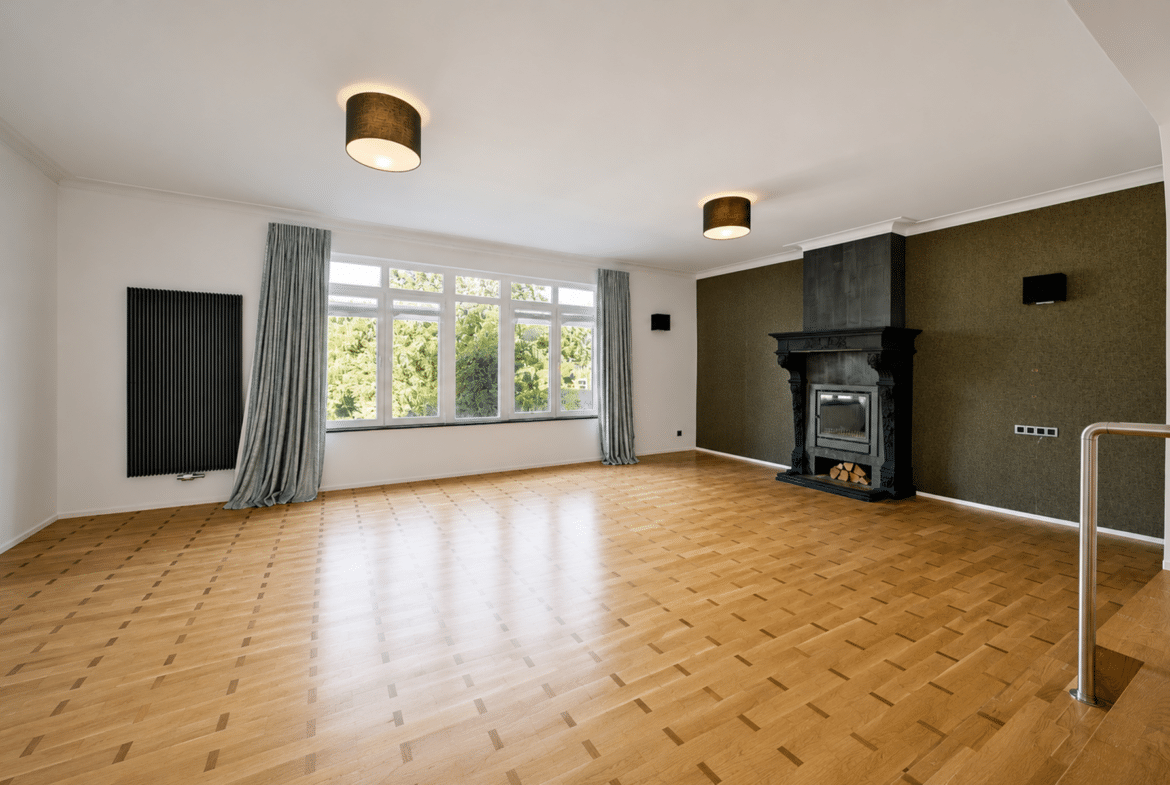Bright living room with large windows, light parquet floor, and an ornate black fireplace on an olive accent wall.