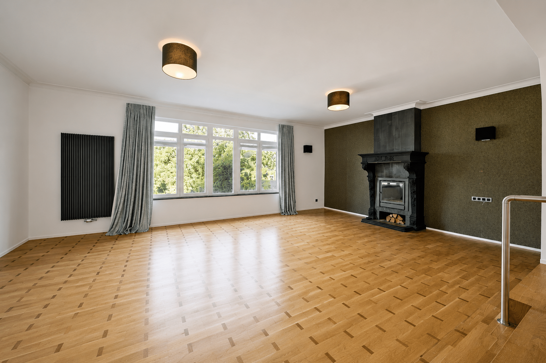 Wijze foto 11 Lichte woonkamer met grote ramen, lichte parketvloer en een sierlijke zwarte open haard op een olijfkleurige accentmuur.