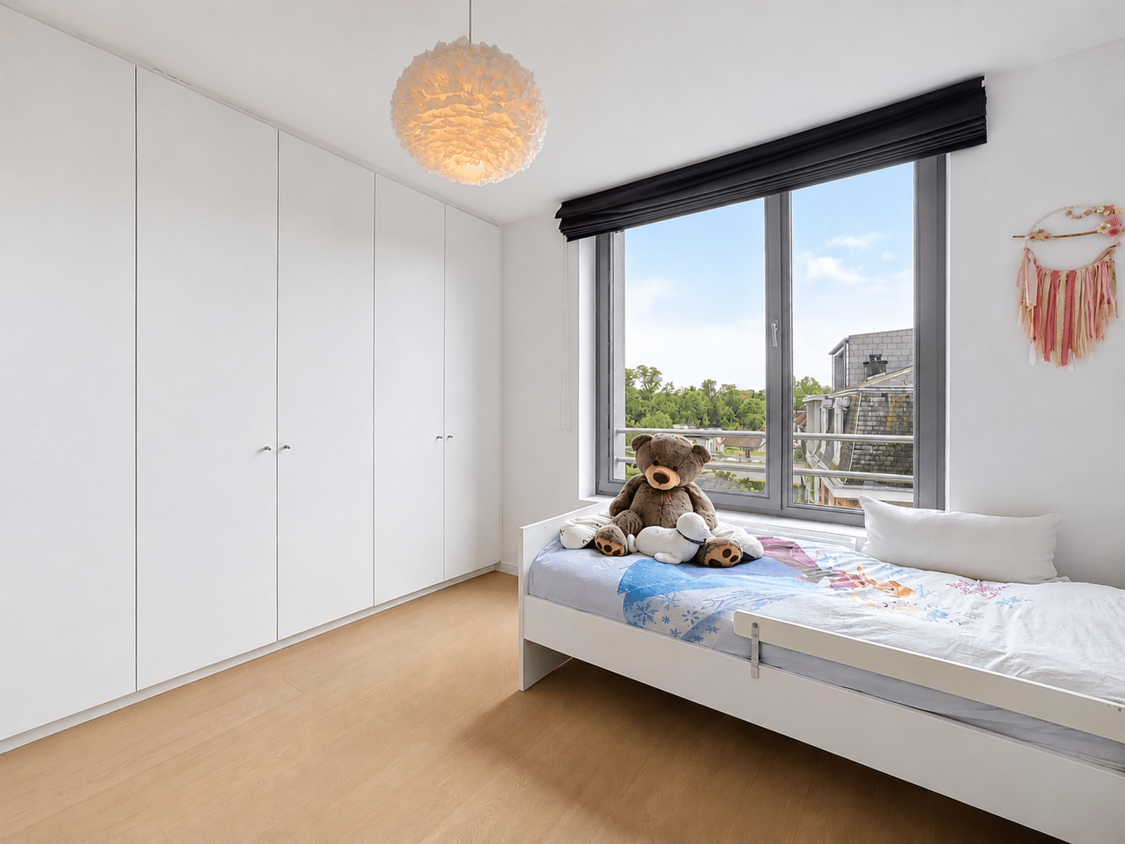 Chambre d’Enfant Lumineuse – You Real Estate Agency Bright children’s bedroom with white built-in wardrobes along the wall, a bed by a large window, and a teddy bear on the bed, a round pendant light overhead, and a decorative tassel wall hanging.