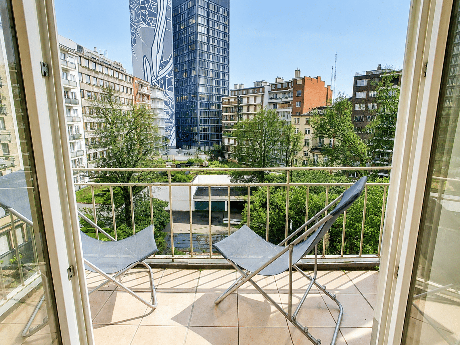 whise photo 12 View from a balcony with two metal chairs overlooking a city courtyard and trees under a clear blue sky in a residential area.