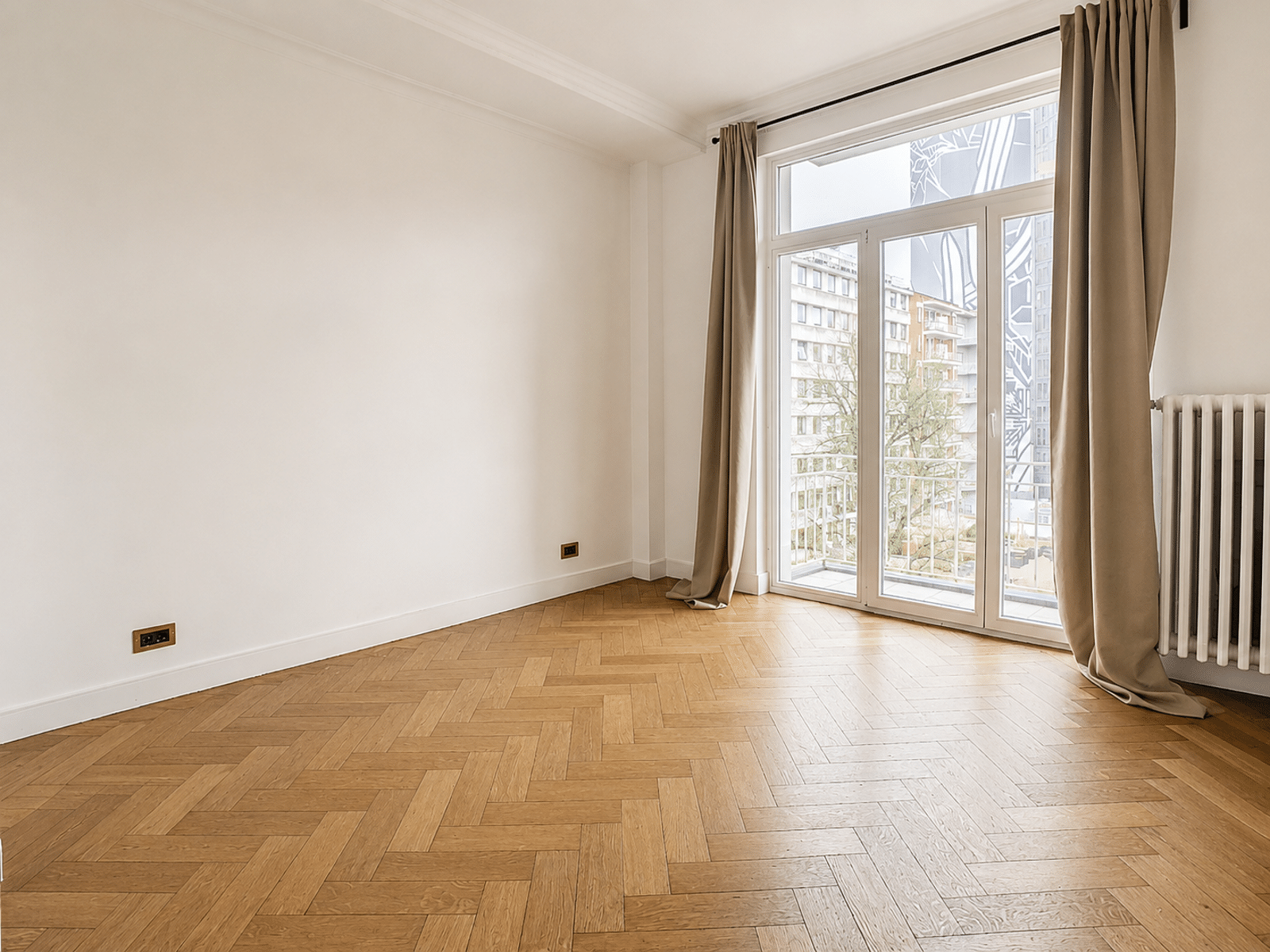whise photo 10 Empty bright room with large glass doors to a balcony, beige curtains, and a herringbone wood floor; radiator on the right at the edge of the wall