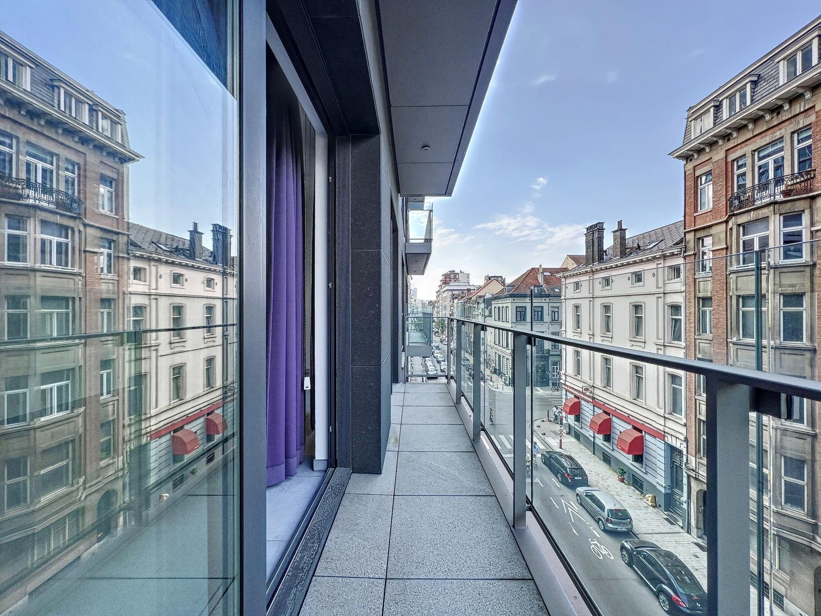 whise photo 8 View from a narrow balcony with glass railing along a modern building, overlooking a city street lined with historic brick buildings under a blue sky
