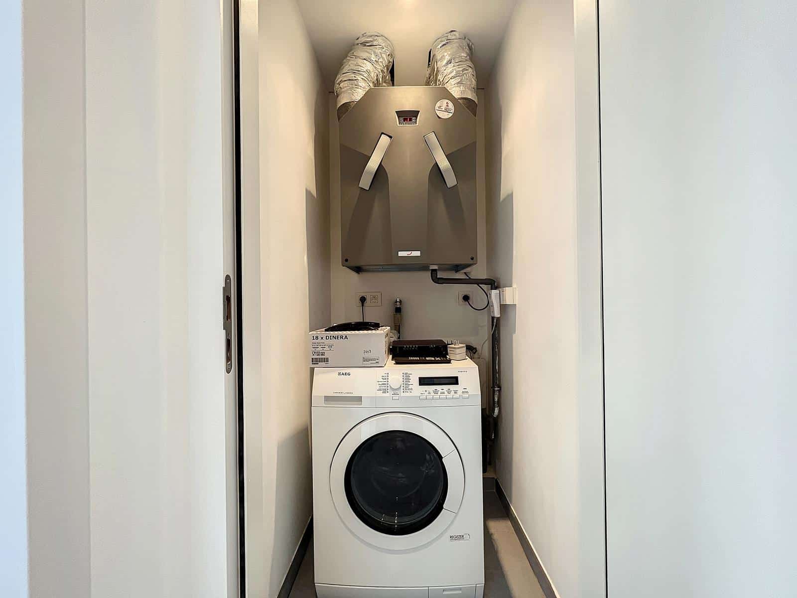 whise photo 6 Small utility closet with a front-loading washing machine and a ceiling-mounted HVAC unit above, with ductwork nearby.
