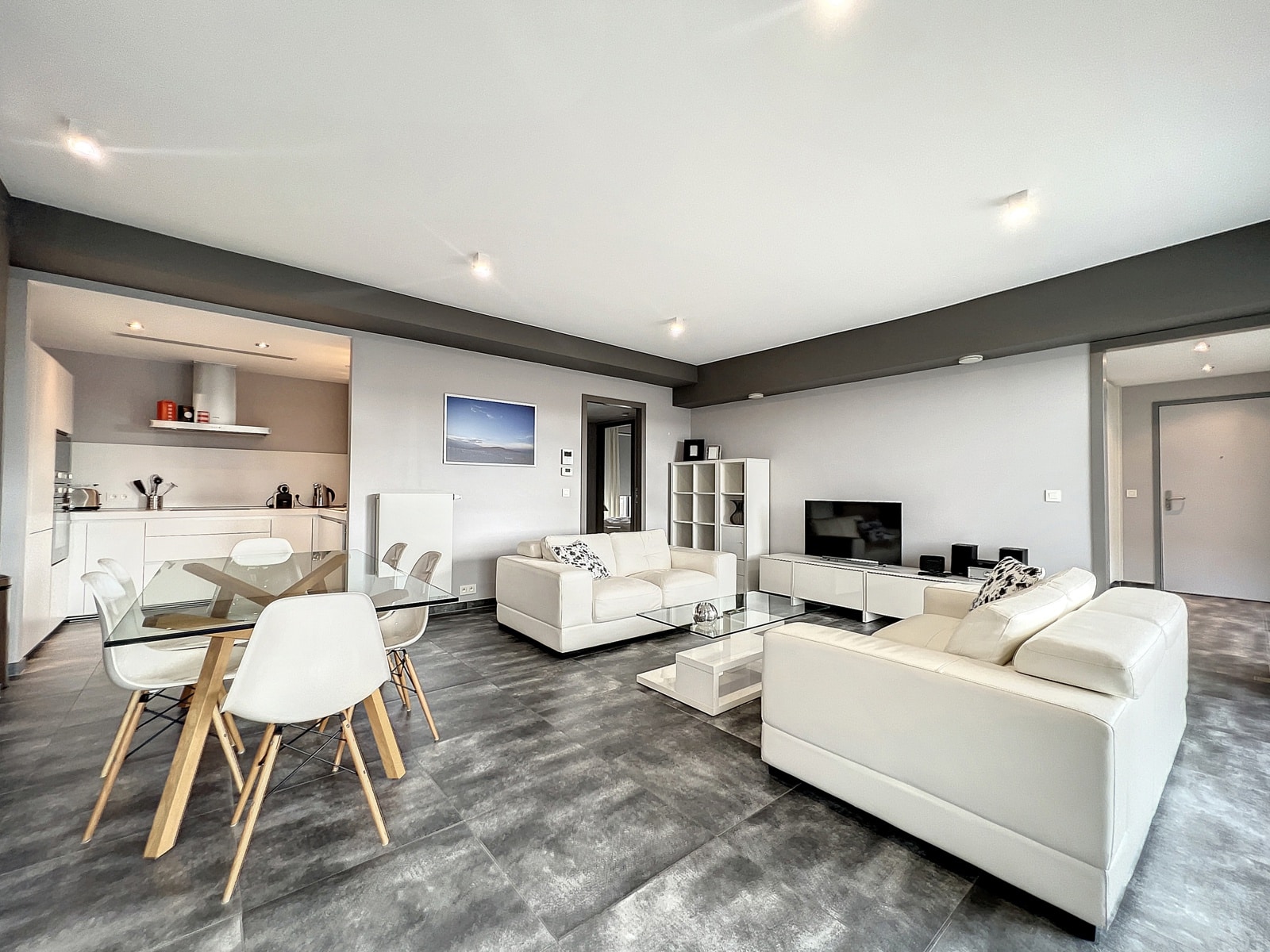 whise photo 2 Bright open-plan living area with white leather sofas, glass coffee table, and a wall-mounted TV unit in a modern gray space.