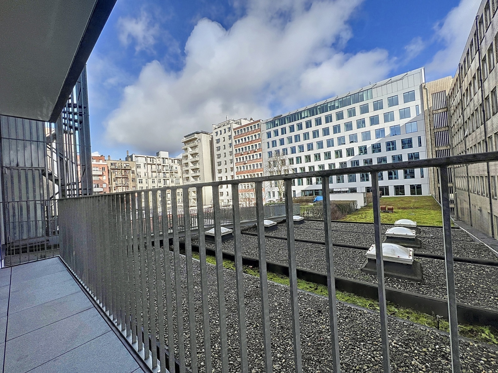 whise photo 11 Balcony railing with urban rooftops and modern buildings in the background under a blue sky with clouds.