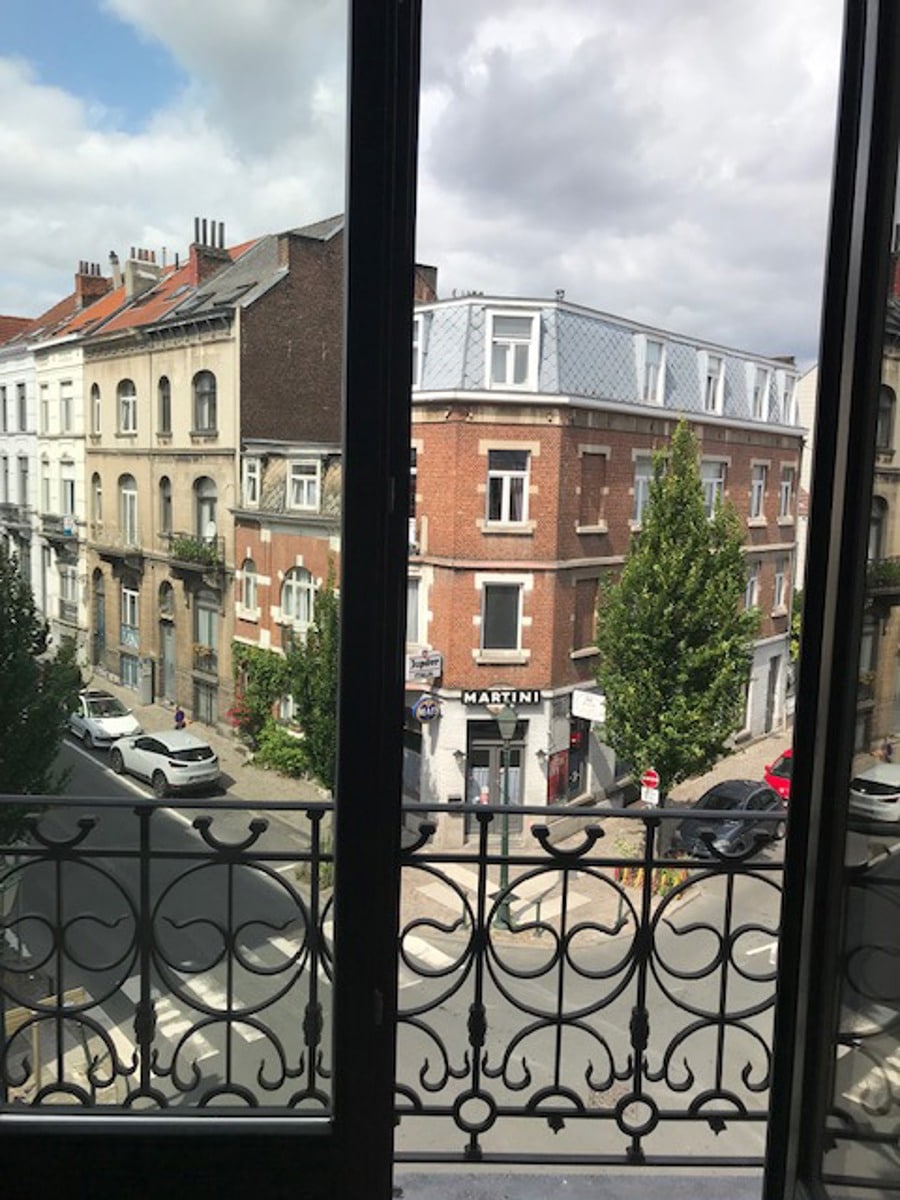 whise photo 10 View from a window onto a European street with brick buildings, a Martini storefront, parked cars, and a decorative iron balcony railing in the foreground.