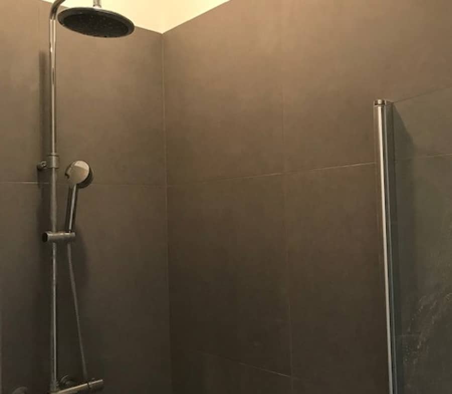 Shower area with a large round rain showerhead and a handheld shower on gray tiled walls, glass door on the right.
