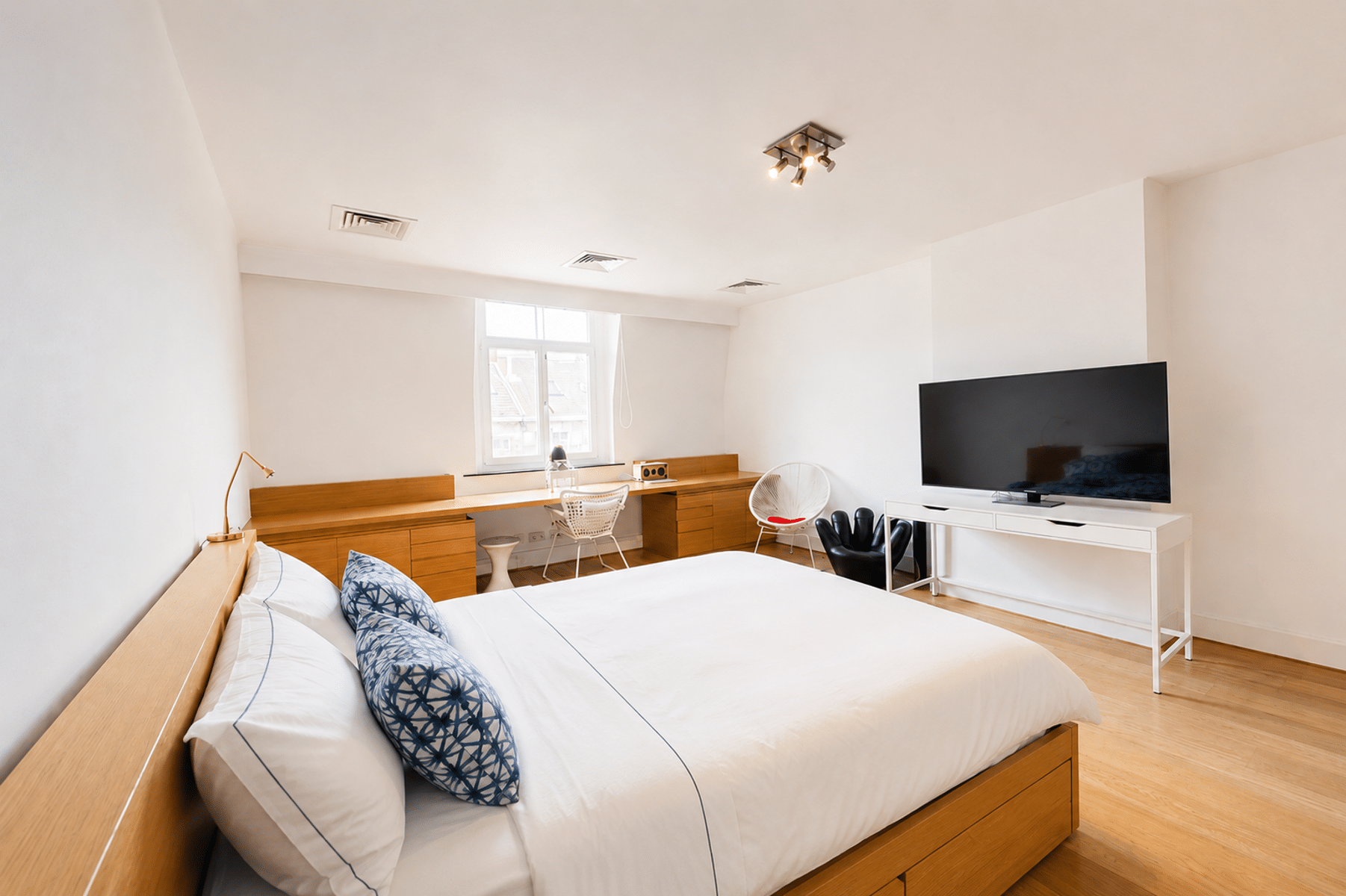 whise photo 16 Bright minimalist bedroom with a wooden platform bed, white linens, and blue patterned pillows; desk and chair by a window in the background.