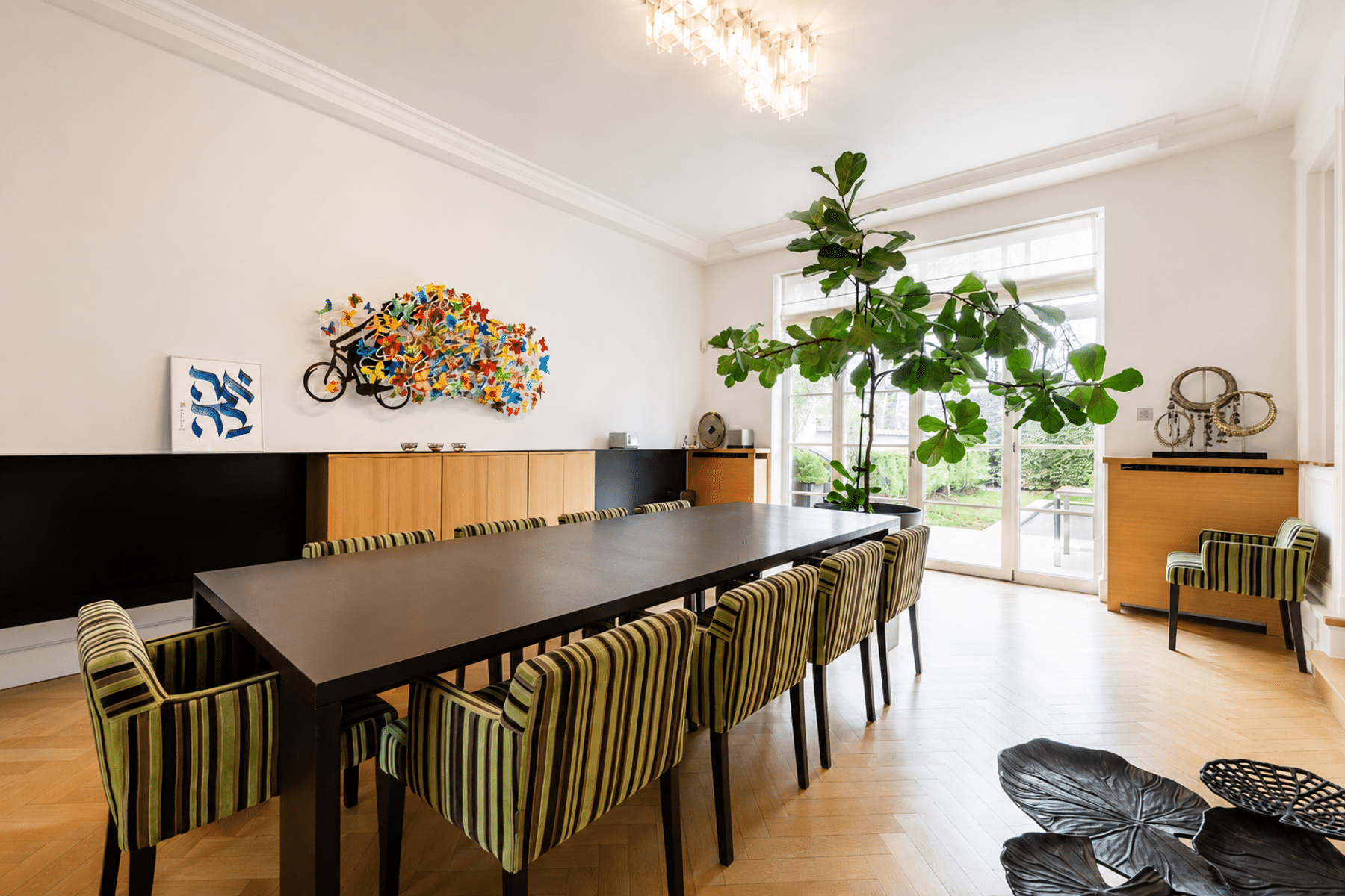 whise photo 7 Modern dining room with a long dark table, striped green chairs, wall art, and a large leafy plant by glass doors.