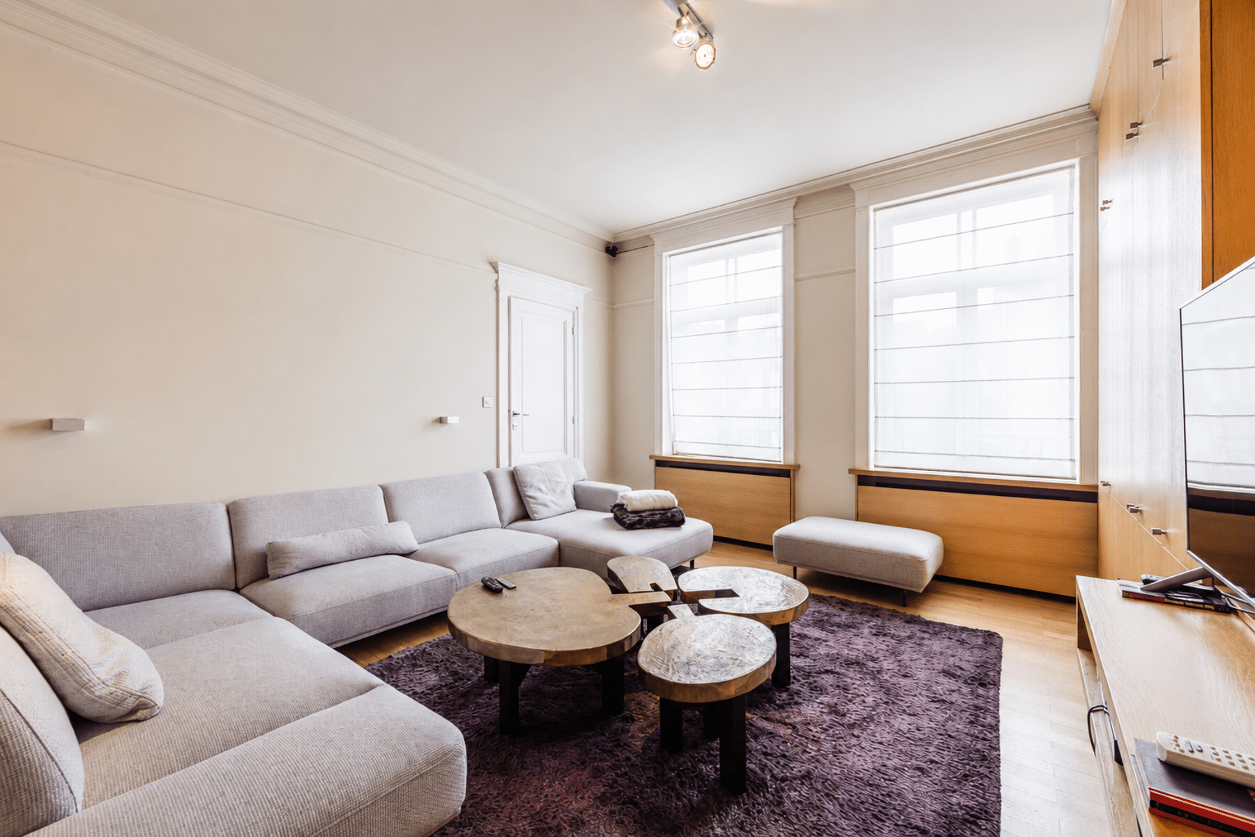whise photo 15 Bright living room with a light gray sectional sofa, purple rug, and small round wooden coffee tables by large windows with blinds.