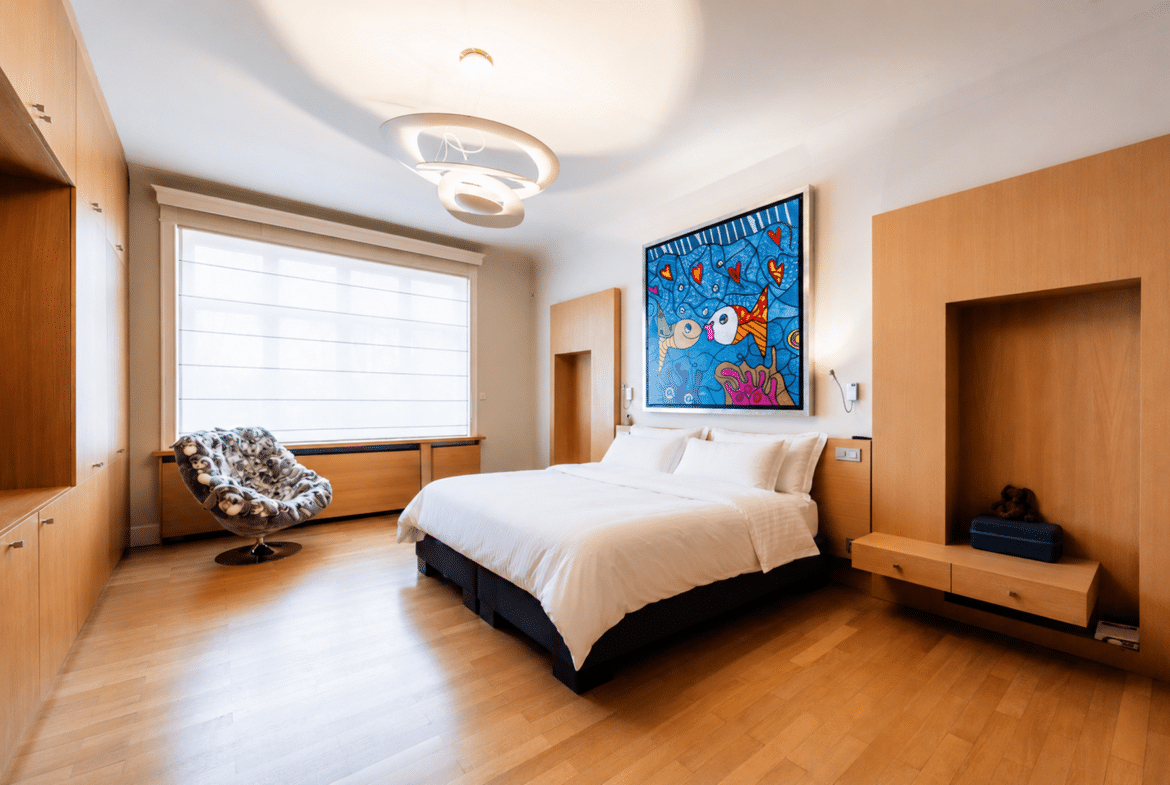 Modern bedroom with white bedding, light wood built-ins, and a colorful framed painting above the bed.