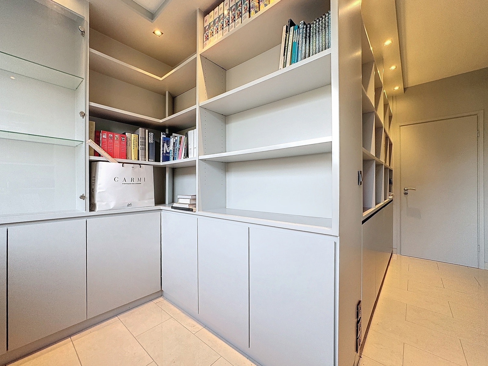 whise photo 16 Corner view of a white built-in shelving unit filled with books, a CARMÍ shopping bag, and lower cabinets along a hallway.