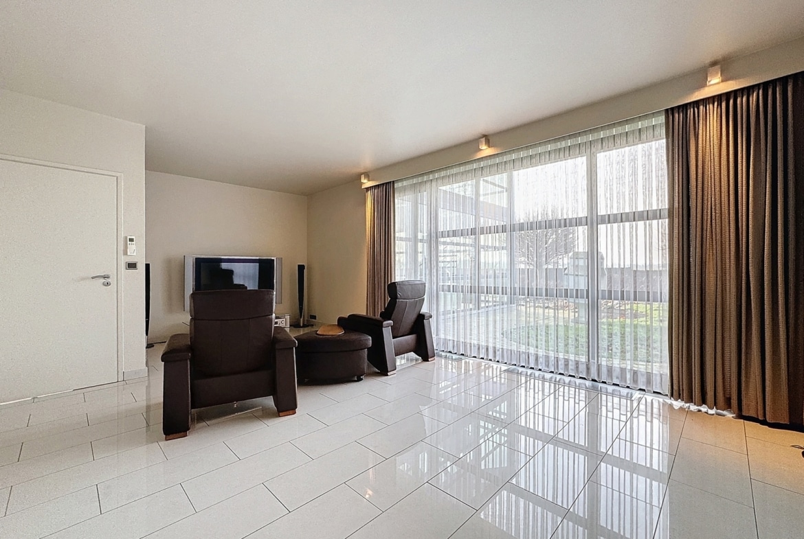 A bright living room with white tile flooring, two brown leather recliners facing a flat-screen TV, and large windows with sheer curtains.