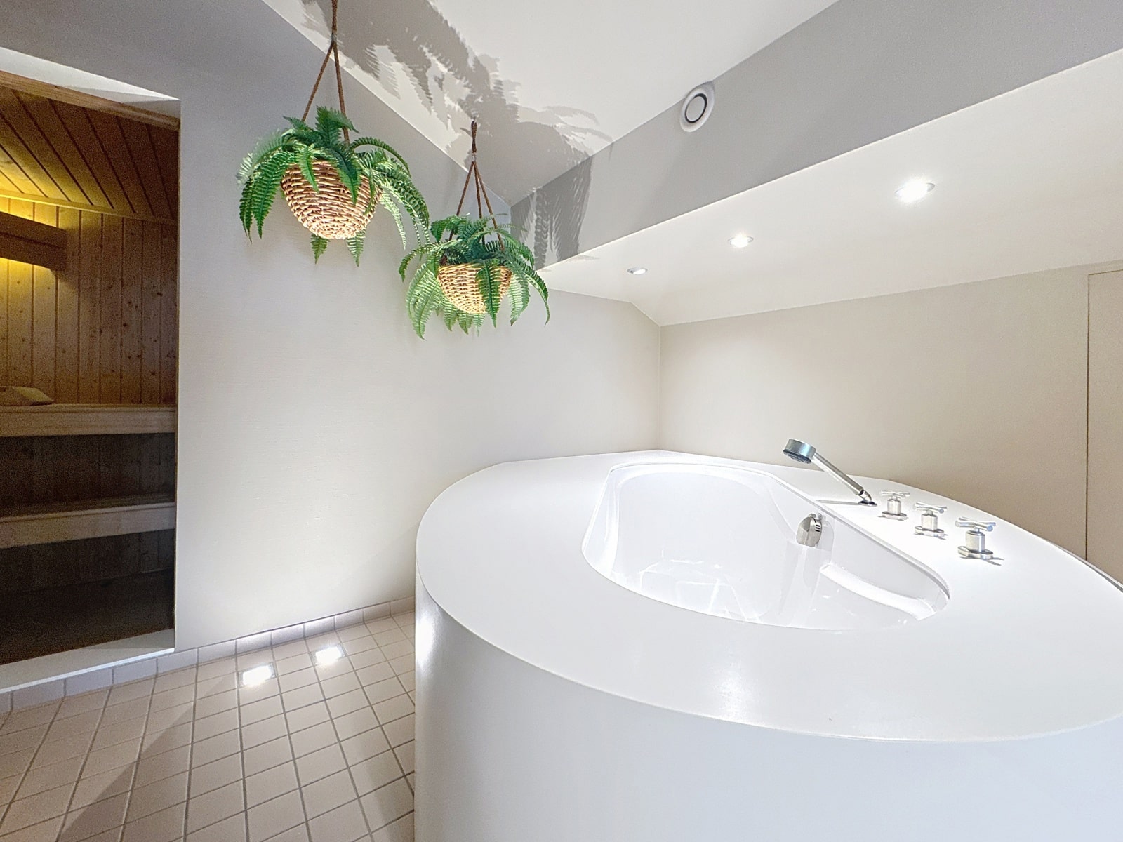 whise photo 23 Modern white freestanding tub with built-in faucet and controls in a bright spa-like room; hanging ferns in baskets above and a sauna door on the left.