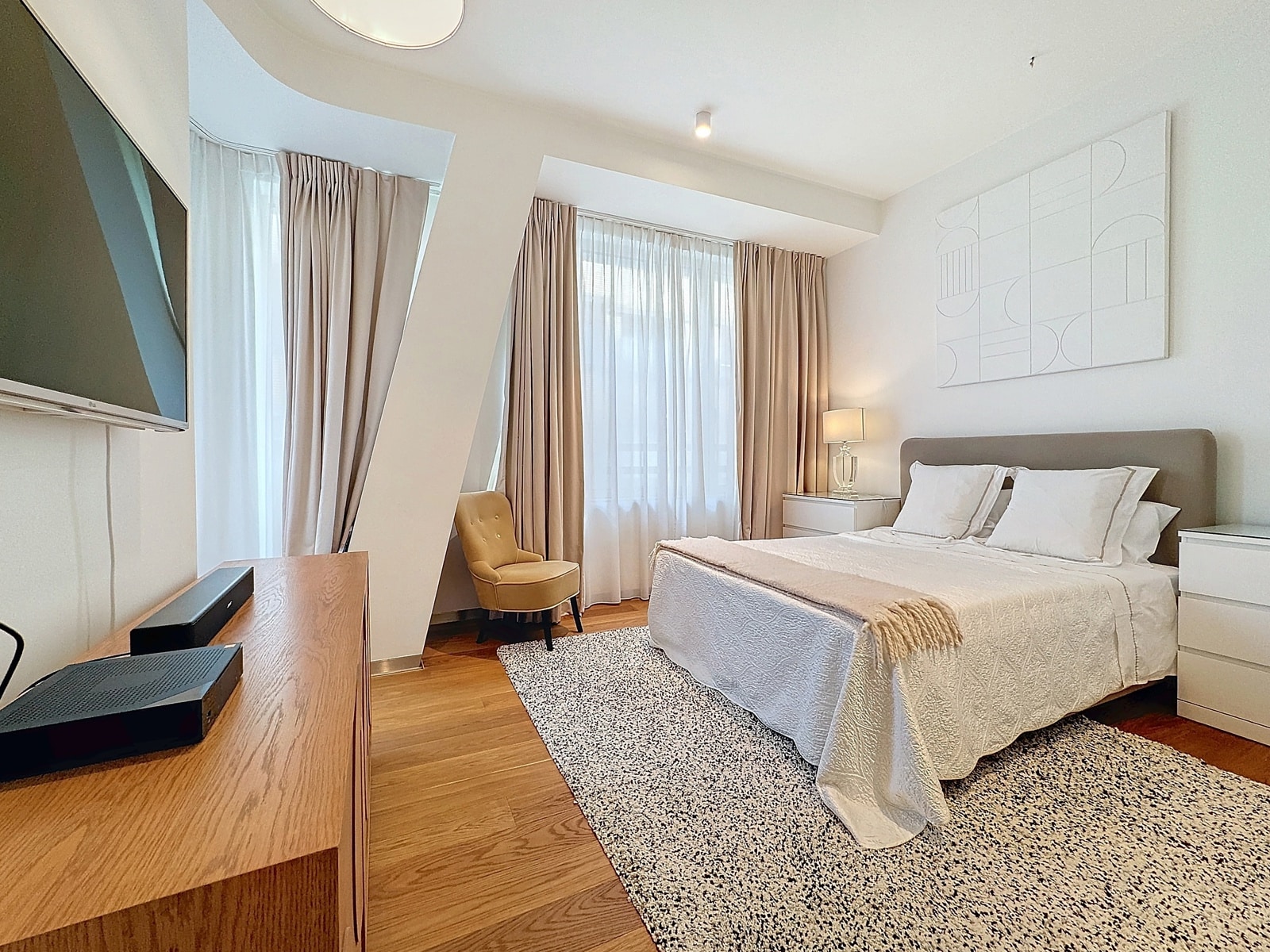 whise photo 8 Bright hotel bedroom with a king bed, white bedding, and beige headboard; front desk area and large windows with sheer white and beige curtains in the background.