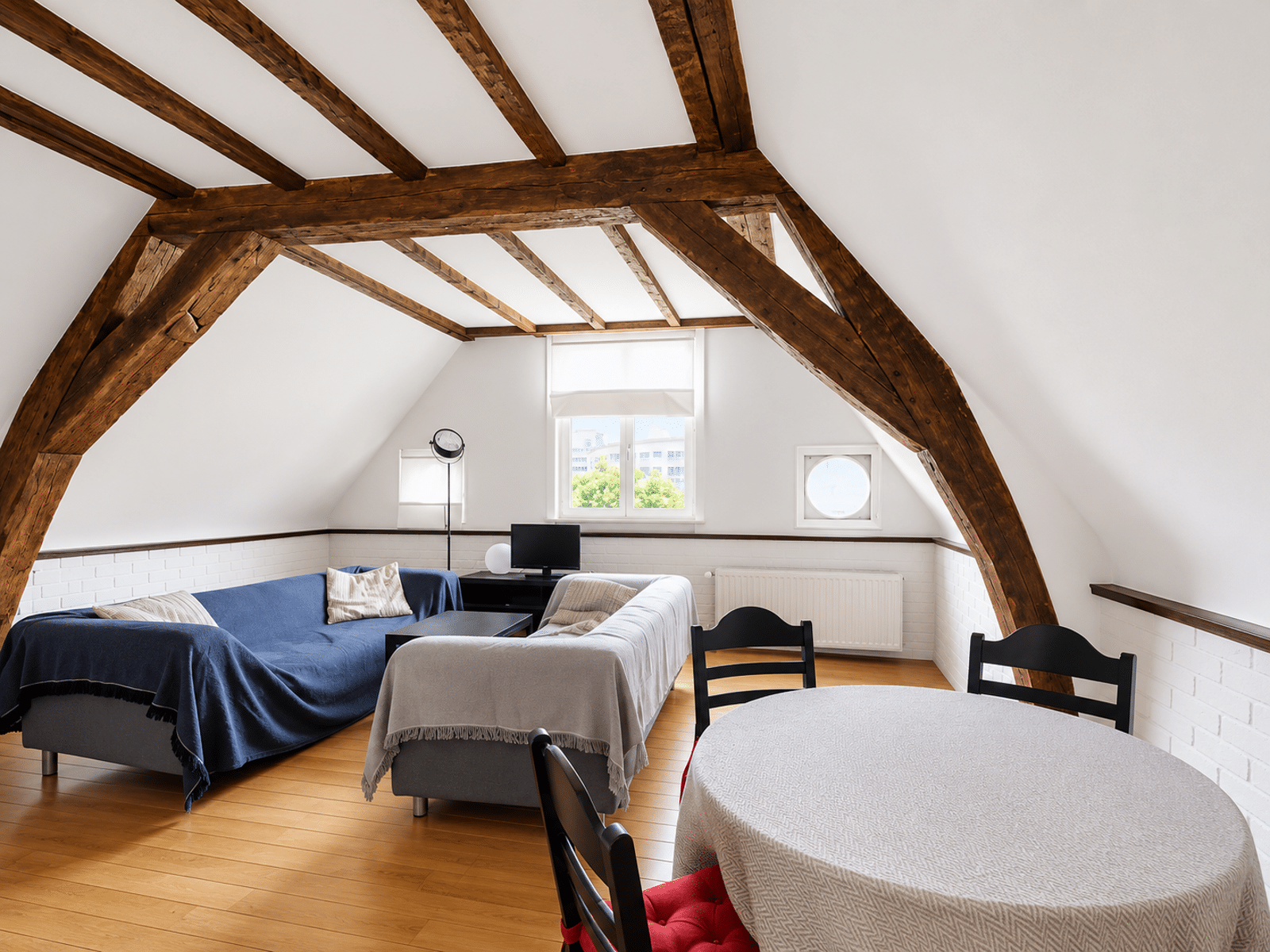 whise photo 2 Attic living room with dark exposed wooden beams, white walls, two couches with throws, and a round table by the window wall.