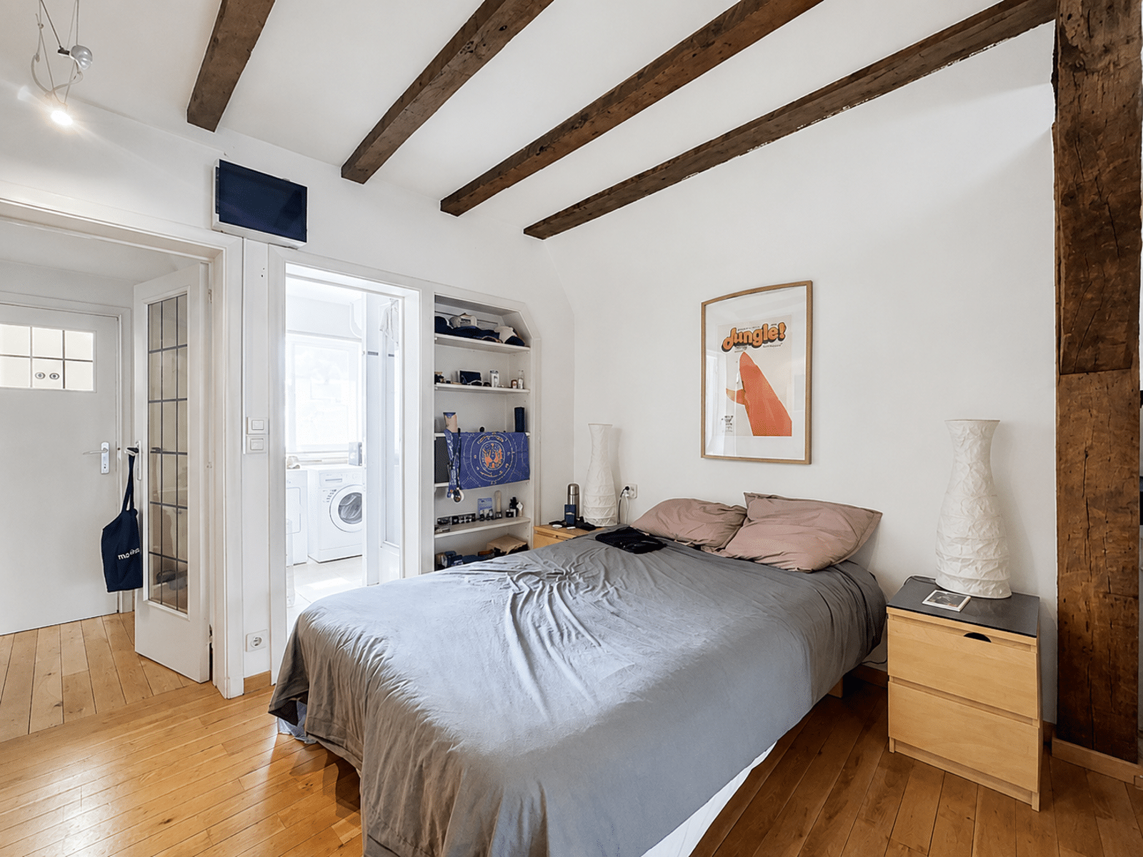 Chambre avec Poutres Apparentes – You Real Estate Agency Bedroom with a gray duvet and pink pillows, exposed wooden ceiling beams, and a framed Jungle! poster on the wall next to a bedside table with a tall white lamp.