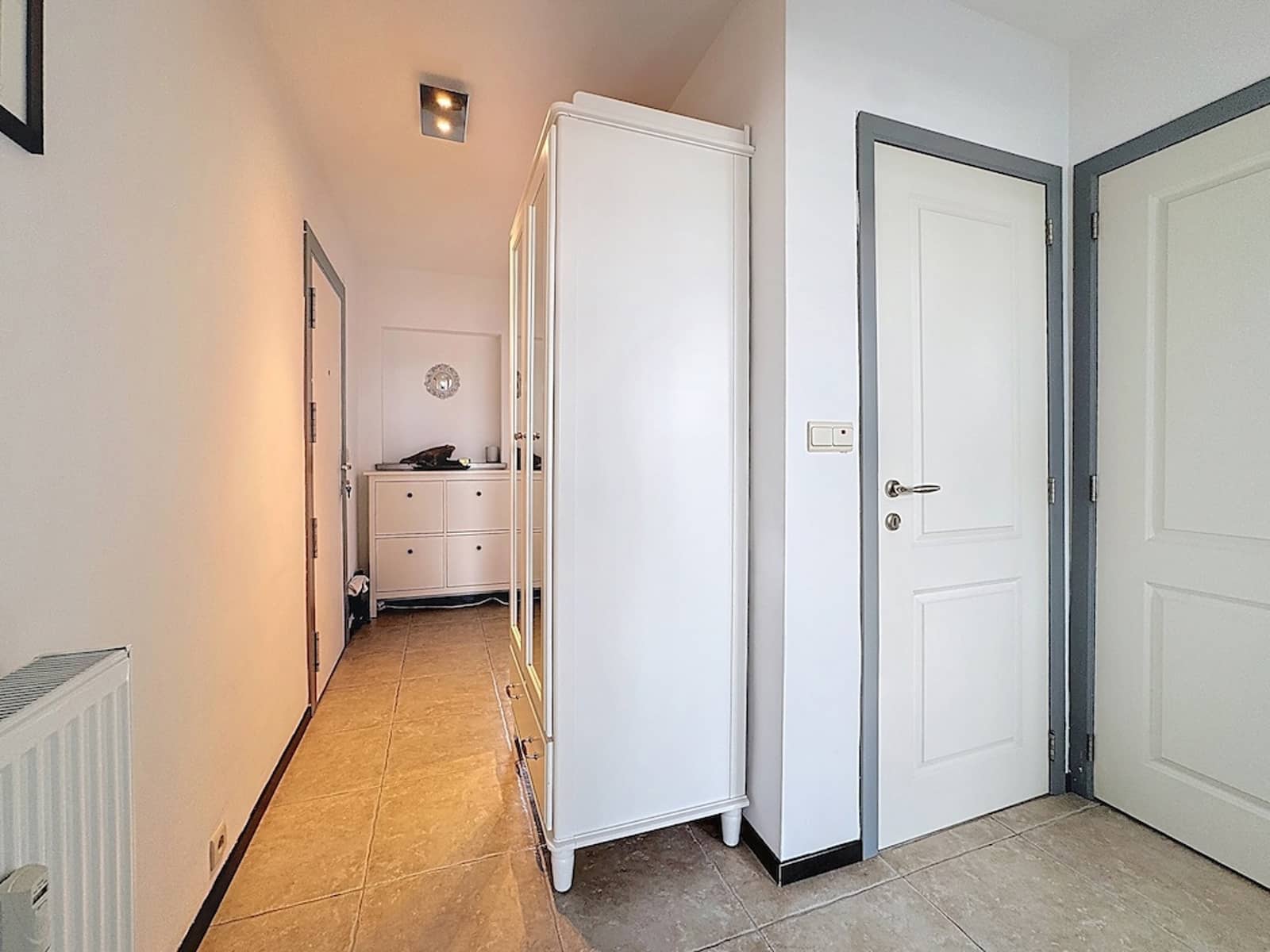 whise photo 11 A long hallway with a white wardrobe on the right, white doors at the end, and a cream-colored dresser visible at the far end; warm overhead lighting and a tiled floor.